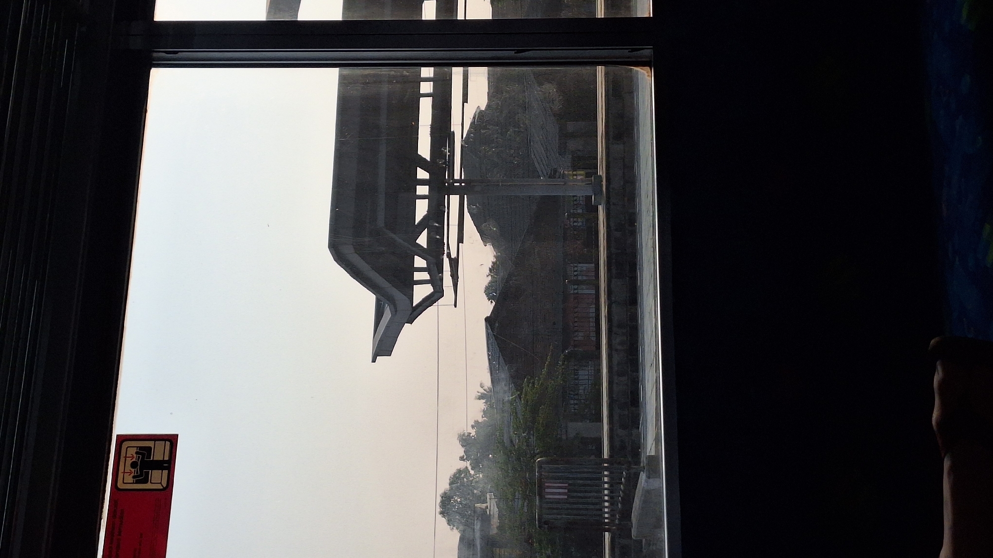 This media file appears to be a photograph taken through a window, possibly on a train or bus, looking out at an industrial or urban landscape. The image is rotated 90 degrees clockwise.
The primary subject in view is an elevated structure, possibly an overpass or part of a building, with a complex framework of beams and supports. Below this structure, a dense collection of buildings with brick facades and roofs are visible, interspersed with trees and greenery. The overall impression of the scene is one of a developed, possibly industrial area, with signs of aging or disuse in some of the structures.
There are no people clearly visible in the photograph. Objects present include the window frame, a red sticker with a graphic symbol on the lower left portion of the glass, and parts of the industrial landscape. The graphic symbol on the sticker appears to represent a card insertion mechanism, possibly indicating the location of a card reader or slot.
The sky is bright and hazy, suggesting it could be daytime, possibly late afternoon given the warm, diffused light. The weather appears to be clear but with some atmospheric haze. Location cues are limited to the general industrial aesthetic and the provided context of Cikarang, Indonesia. There are no discernible emotions conveyed by the image, as it focuses on the external environment.
There is text on the red sticker. The graphic symbol is black and white with red accents. The red sticker itself has some red markings and potentially white text, though it is not legible at this resolution.