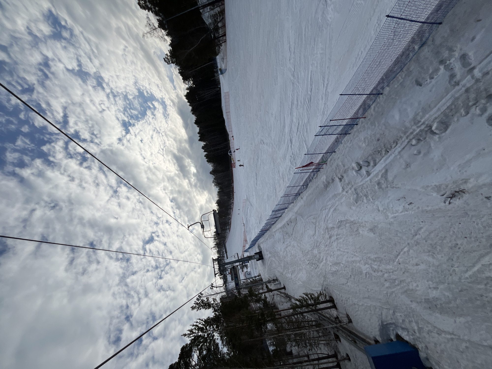 The image depicts a snow-covered ski resort landscape, observed from an elevated position, likely a chairlift. The sky above is partially cloudy, featuring a mix of white stratocumulus clouds and visible patches of blue sky. Three dark chairlift cables run diagonally across the upper portion of the frame.
In the foreground, a snow-covered track runs parallel to a fence line. To the left of this track, a dense row of pine trees and some bare deciduous trees ascend, partially obscuring a blue, rectangular object at the bottom-left. A prominent metal chairlift tower, supporting multiple empty chairlift seats, stands mid-ground on the left side of the frame.
To the right of the track and chairlift tower lies a wide, groomed ski slope covered in white snow, displaying various ski tracks. Several individuals, identified as skiers, are visible as small figures on this slope; some appear to be actively descending, while others are static. The ski slope is delineated from the track by a low fence constructed from blue mesh netting, supported by vertical posts painted alternating red and blue. Further up the slope, orange and white safety netting or windbreaks are present.
The entire right side and background of the scene are bordered by a dense forest of dark, tall trees. The snow on the ground immediately below the viewpoint appears compacted, with visible footprints and some areas where the ground or ice is exposed. No specific city or location details are discernible beyond a generic ski resort environment.