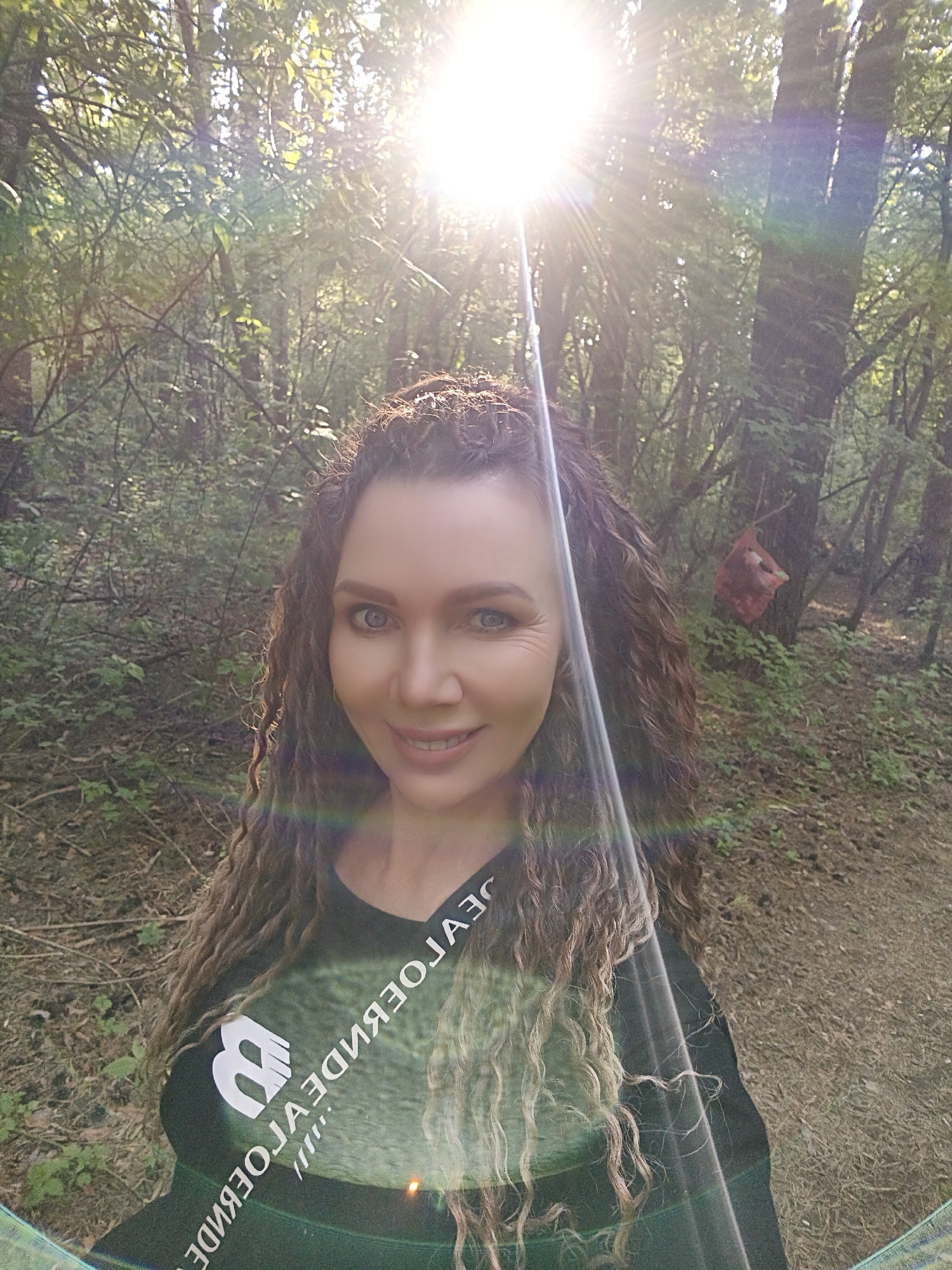 The image is a selfie of a woman in a wooded area on a sunny day in Unknown, Unknown. The woman is fair-skinned with curly brown and blonde hair. She has blue eyes and is smiling. She is wearing a black t-shirt with a white logo and reversed white text. The trees are mostly green and brown, with some sunlight filtering through the leaves. A red object is suspended from a tree branch in the background to the right. The ground appears to be covered in leaf litter.