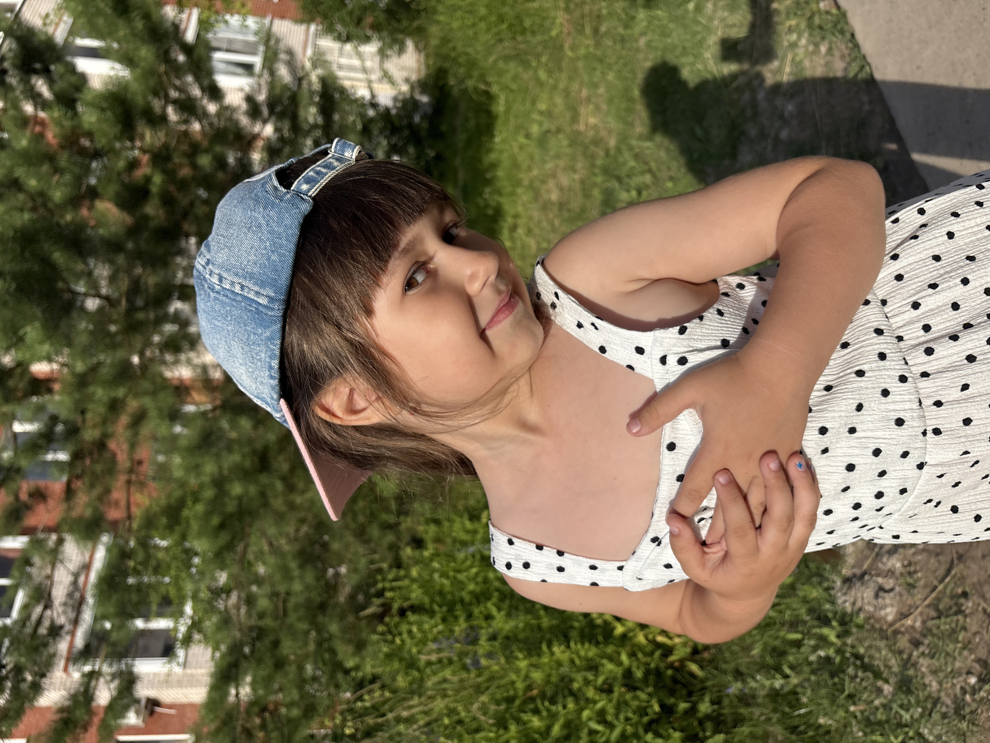 A young girl wearing a denim cap and a white dress with black polka dots is posing for a photo outdoors. She is looking towards the camera with a slight smile and her arms are crossed over her chest. Her hair is brown and she has bangs. The background shows a grassy area with some trees and buildings in the distance, suggesting a residential area or park. The lighting indicates it is daytime, possibly late afternoon due to the shadows. The overall mood seems relaxed and playful. There is no visible text in the image.