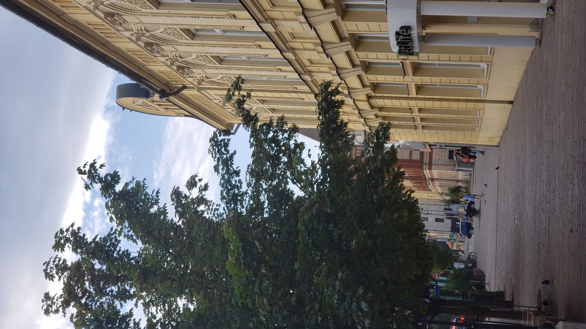 The image depicts a street scene in Prešov, Slovakia, likely during daytime given the lighting conditions. On the right side of the frame is a light yellow building with ornate architectural details and multiple windows. A sign on the building reads "PentaLife". In the foreground, a tree with dark green foliage stands on the left side of a paved walkway. The walkway is made of brick-like pavers and is lined with trees and benches. Further down the street, several people are visible. A blue car is parked near a statue, and two women are standing in the street. A man in a dark jacket is walking on the walkway, and another man is partially visible near a red truck in the background. The sky is partially cloudy with some blue sky visible.