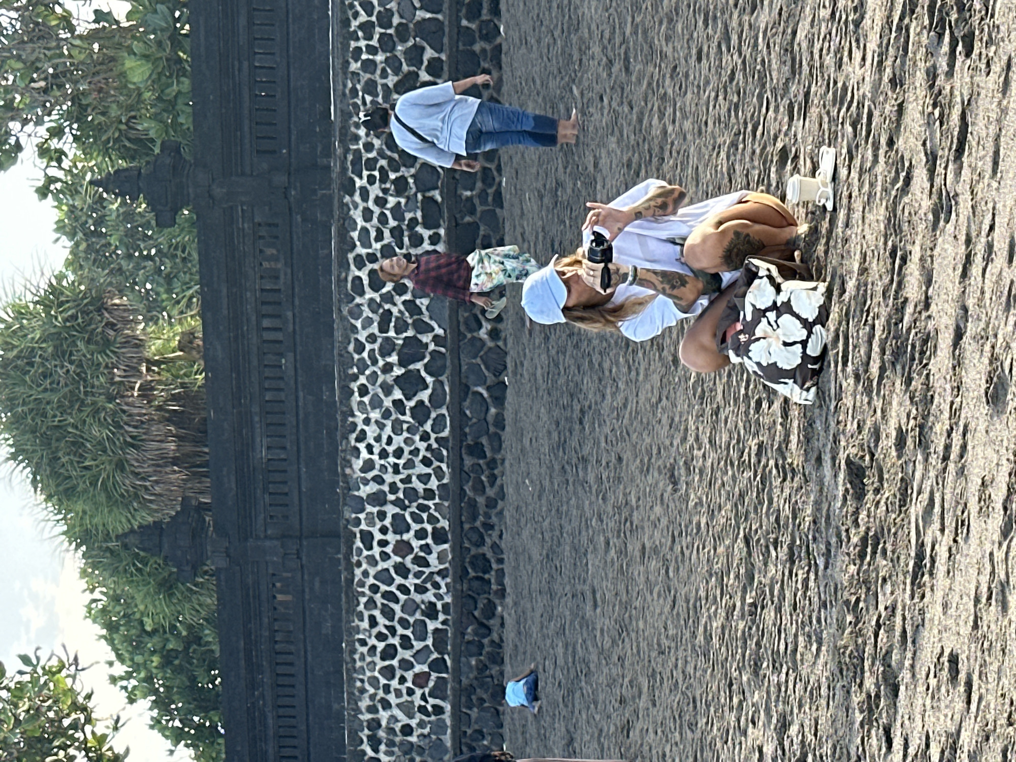 In North Kuta, Indonesia, a beach scene unfolds. In the foreground, a person wearing a white t-shirt and a light blue baseball cap crouches on the sand, holding a camera to their eye and appearing to take a photo. They have numerous tattoos visible on their arms and legs. Beside them sits a patterned tote bag and a small white cup with a saucer and flip-flops. In the middle ground, a person in a red and black plaid shirt and a floral skirt walks away from the camera, holding a pair of flip-flops. To their right, another person wearing a light blue long-sleeved shirt and jeans stands with their back to the camera, facing away from the water. Further back, a child in a blue shirt kneels on the sand. The background features a textured black stone wall topped with a decorative stone fence and lush green foliage. The ground is dark, wet-looking sand.