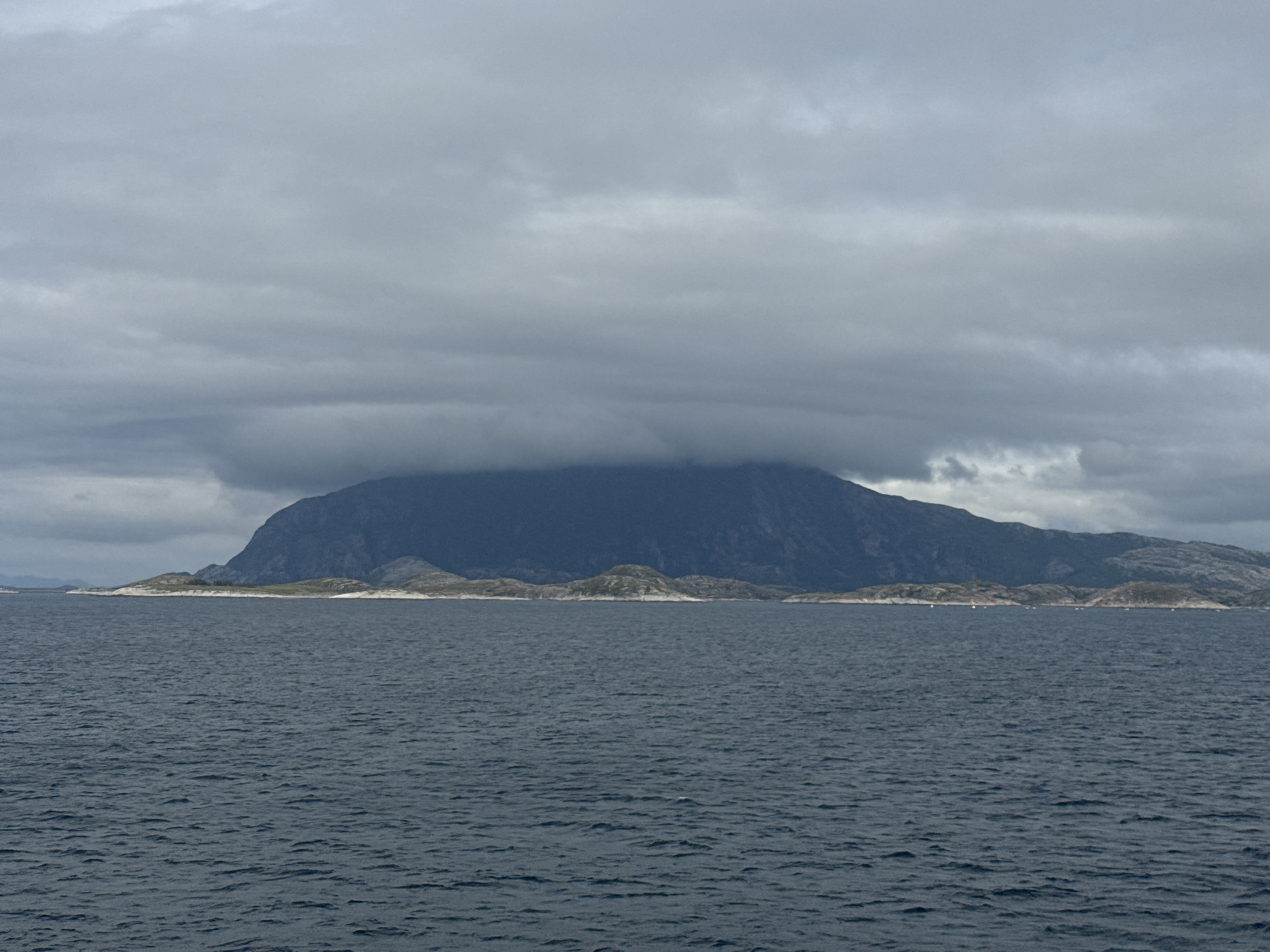 The image shows a wide shot of the sea and a mountainous island under a cloudy sky. The sea is dark blue and choppy. The island, which is part of Onøya, Norway, has rugged, rocky terrain and is partially covered by dark clouds. There are several smaller islands and peninsulas in the foreground, with some vegetation and rocky shores. Several small boats are visible on the water near the islands. The overall scene is overcast and somber.