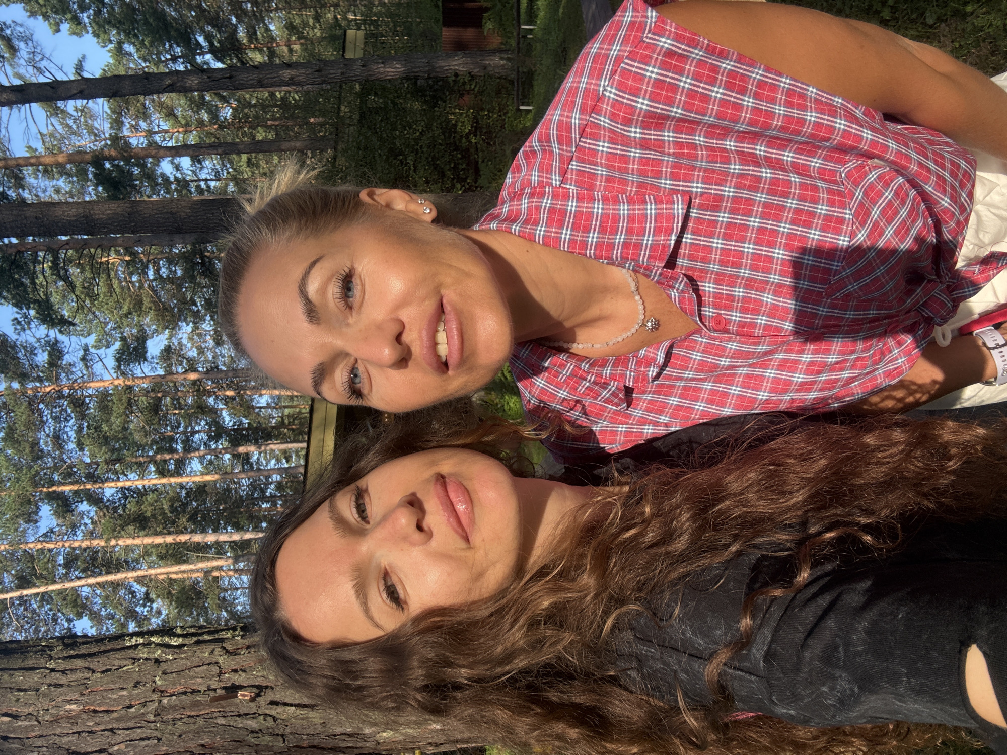 This is a close-up selfie of two women in a forest environment, likely outdoors. The woman on the left has long, wavy brown hair and is wearing a dark top. The woman on the right has blonde hair pulled back and is wearing a red and white plaid sleeveless shirt. Both women are looking towards the camera with smiles. The background consists of tall pine trees with a clear blue sky visible above. Sunlight is casting shadows on the ground and on the subjects. There is no visible indication of the city of Barangol, Russia, in the image.