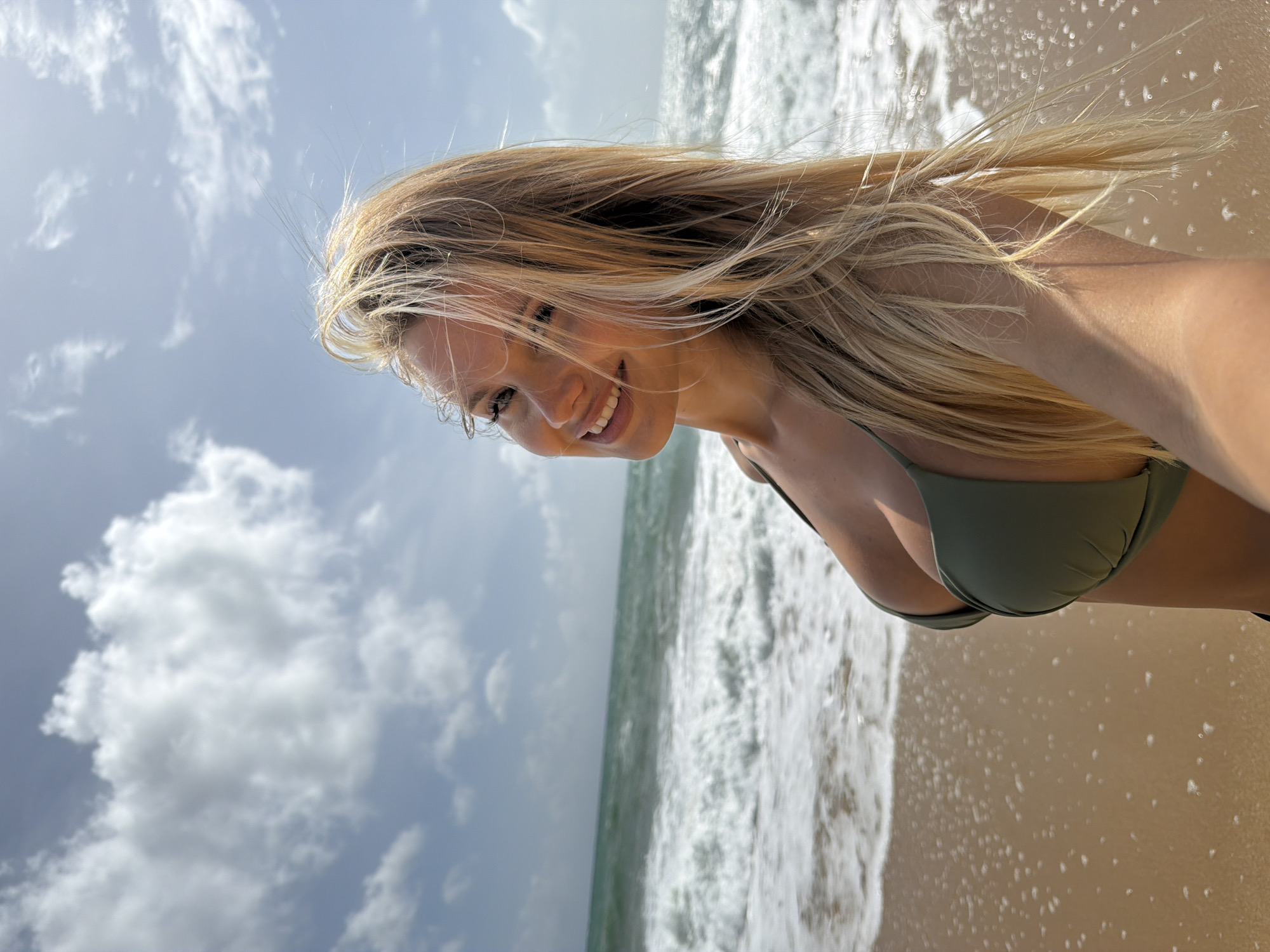 A young Caucasian woman with long blonde hair is taking a selfie on a sandy beach in Mai Khao, Thailand. She is wearing a green bikini top and smiling at the camera. The ocean with waves is behind her, and the sky is partly cloudy. The sun is shining, casting shadows and highlighting her features.