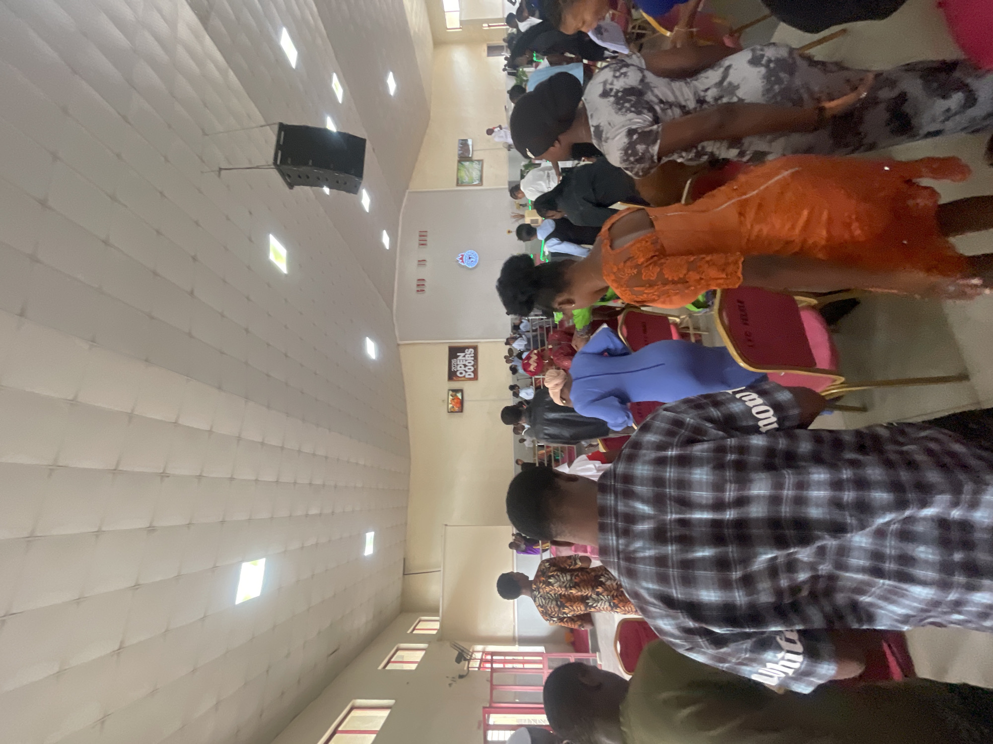 The scene depicts a gathering of people inside a building in Lokoja, Nigeria. The building appears to be a hall or church, with rows of chairs occupied by individuals of varying ages and dressed in diverse attire. Some are seated while others are standing or moving about. The environment is brightly lit, with windows providing natural light. A sound system is suspended from the ceiling. There are signs and posters on the walls, including one that says "God is love" and "Open doors 2023".