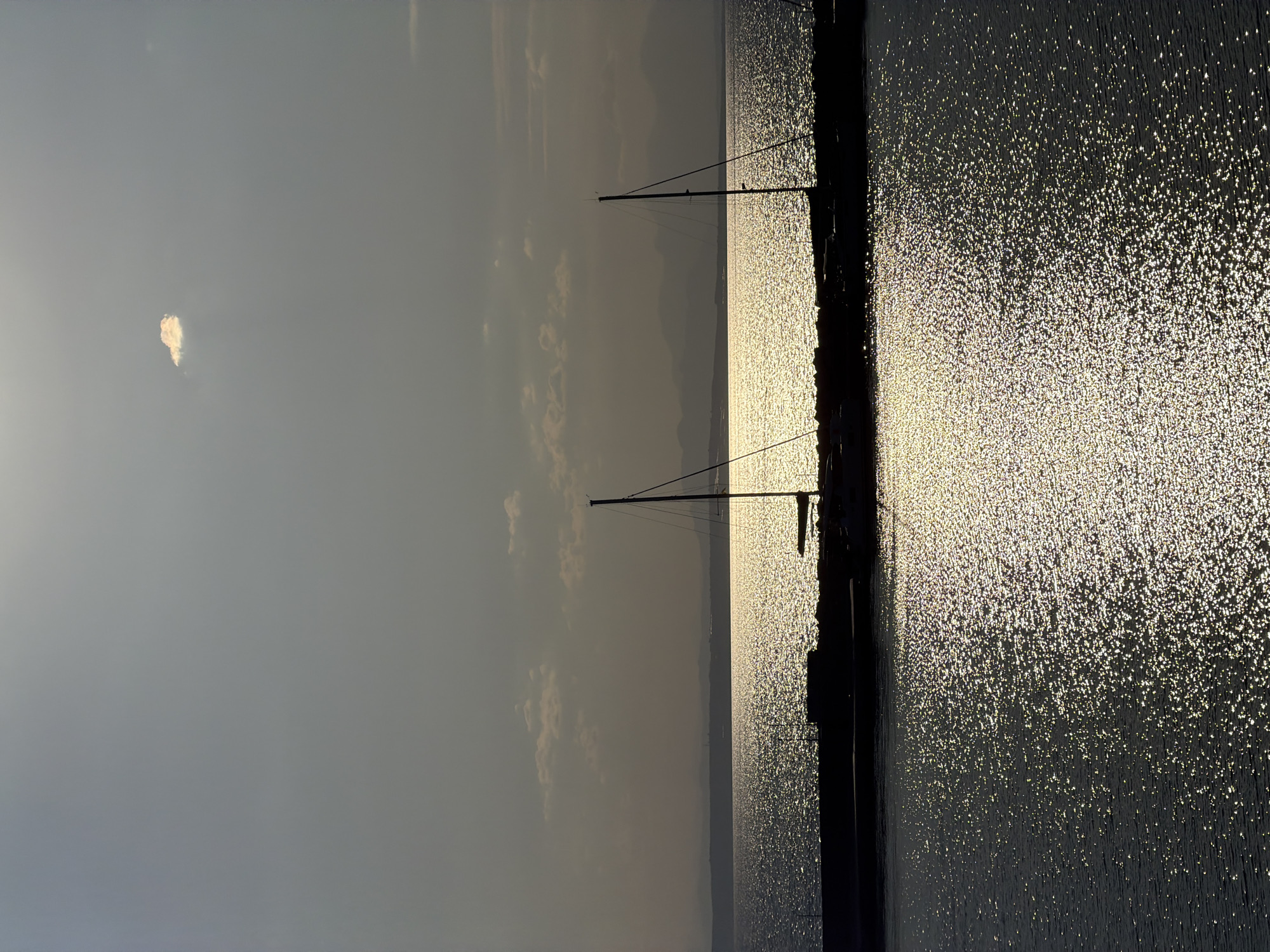 The image shows a view of the Ionian Sea in Zakynthos, Greece, during what appears to be sunset or sunrise. The sun reflects off the water, creating a shimmering effect. Silhouetted against the bright water are two sailboats moored at a dock. In the distance, faint outlines of mountains are visible on the horizon. A single, small cloud is present in the upper portion of the frame. The overall atmosphere is serene and tranquil.