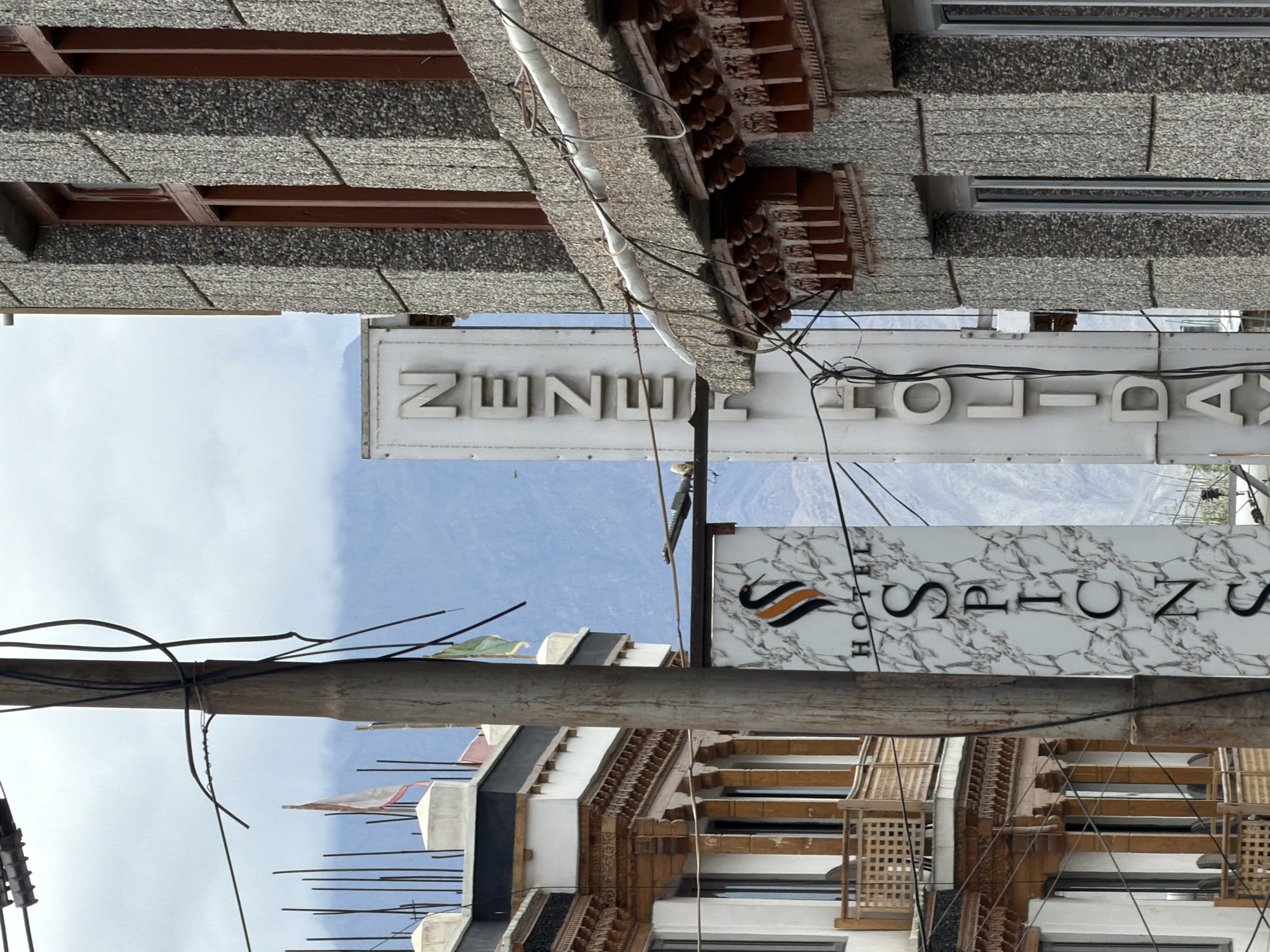 The image shows a street scene in Leh, India, with a focus on the architecture and signage of buildings. In the foreground, a utility pole with numerous wires is prominently featured. To the left, a multi-story building with traditional Ladakhi architectural elements, including ornate wooden balconies and windows, is visible. A sign for "HOTEL SPICNS" with a stylized bird logo is attached to a pole in front of this building. To the right of the utility pole, a vertical sign reads "NENZE HOLIDAY." Behind these structures, a larger building with a textured stone facade and dark wooden window frames is visible. In the background, hazy mountains and a cloudy sky suggest the mountainous terrain surrounding Leh. The scene appears to be a typical street view in the town.