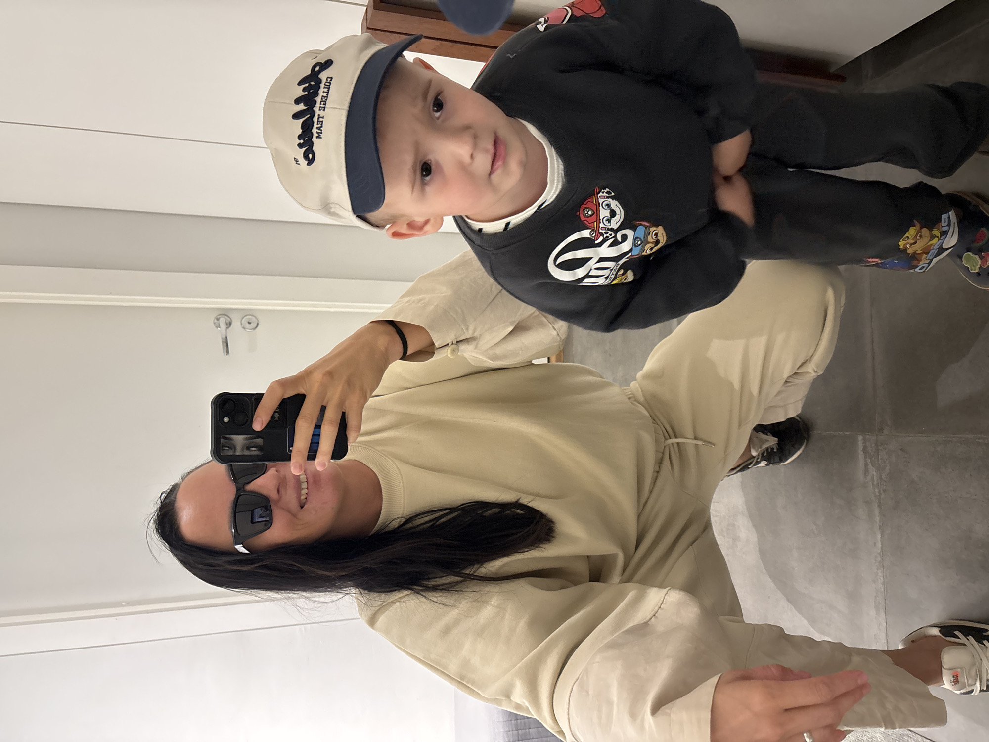A woman and a young boy are posing for a selfie, likely in a bathroom. The woman, wearing sunglasses and a light beige sweatsuit, is crouching and holding up a smartphone to capture the image. The boy, positioned above her, is wearing a cap, a dark sweatshirt with cartoon characters, dark pants, and character-themed shoes. The background includes a door, bathroom fixtures, and tiled flooring. There is no information in the image to indicate that this scene is taking place in Chaikovsky, Russia.
