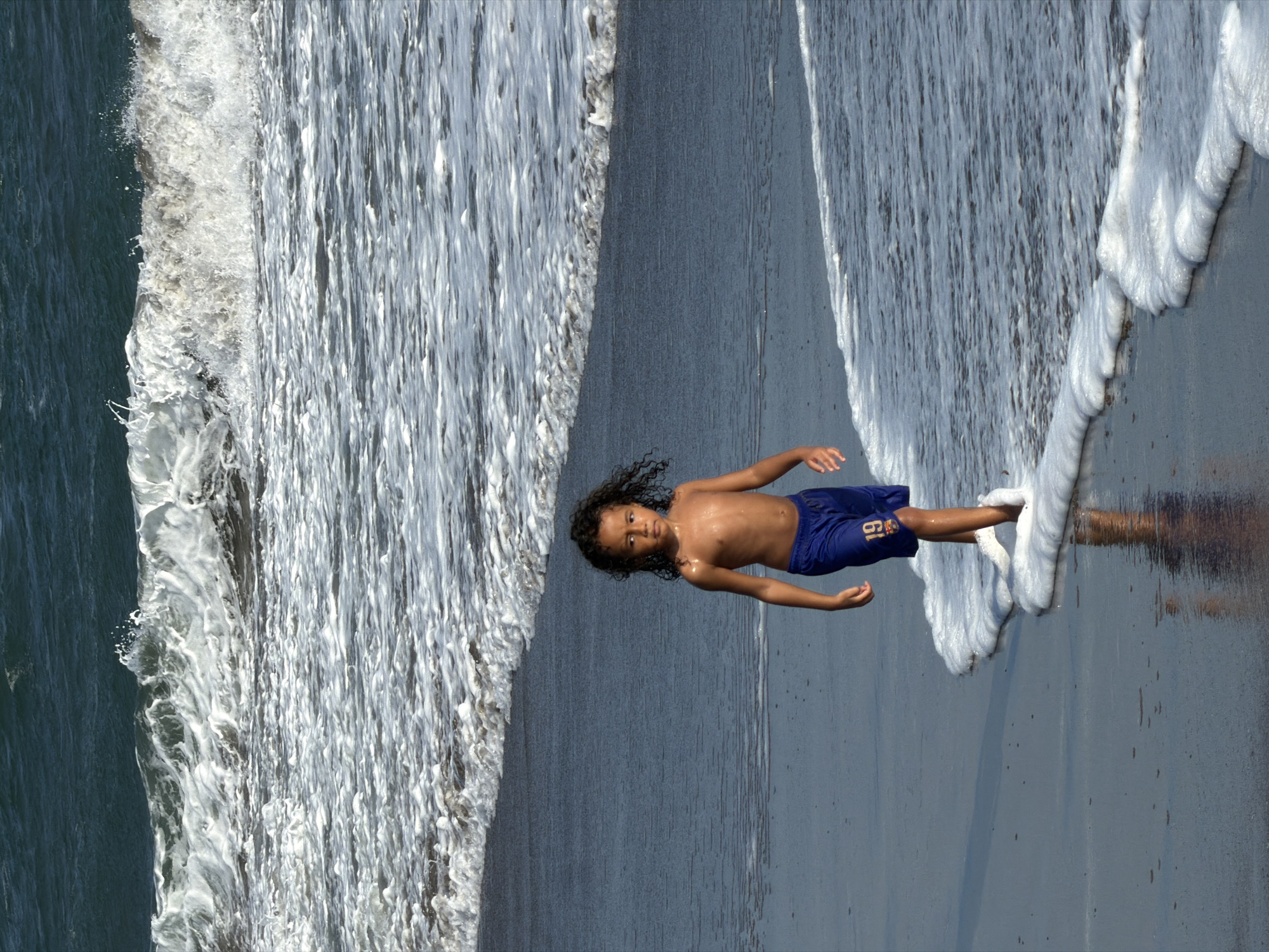 A young boy with curly dark hair stands on a beach, facing the camera with a neutral expression. He is shirtless and wearing blue swim trunks with the number "19" and a logo on the left thigh. The boy is partially submerged in foamy white water, which appears to be a wave breaking or receding on the shore.
The scene is a beach in North Kuta, Indonesia. The water on the left side of the image is dark and deep, with a foamy crest indicative of a breaking wave. The sand or wet ground where the boy stands is dark and reflects the water. The light suggests it is daytime, possibly late afternoon given the warm tones on the boy's skin. The weather appears to be sunny, with the water creating a dynamic and active scene. The composition of the image is unusual, as the scene is rotated 90 degrees clockwise, making the waves appear to be flowing vertically.
