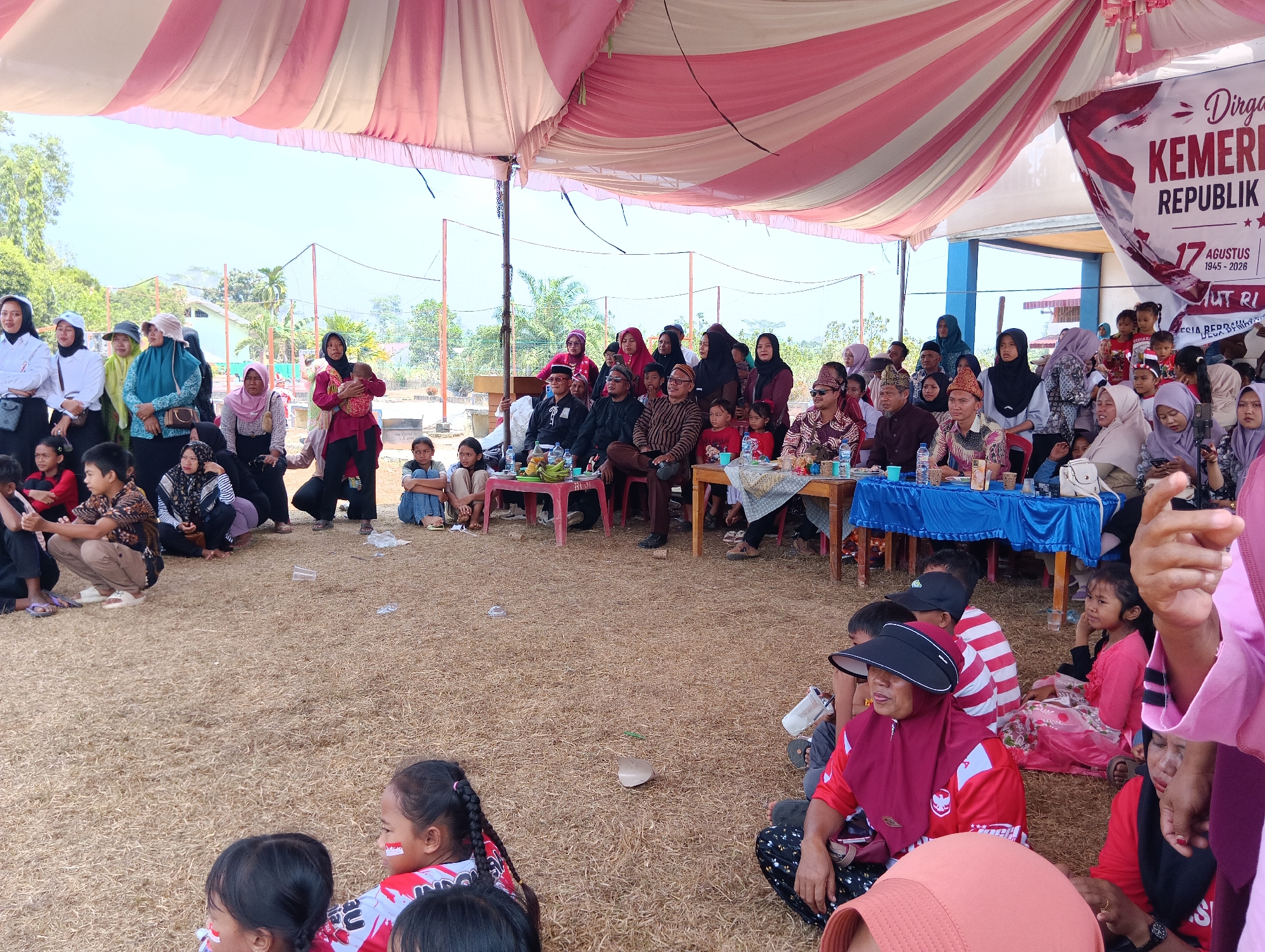 The image depicts an outdoor gathering under a tent with a striped pink and white canopy, possibly in Pematang Jaya, Indonesia, given the context. A large banner reading "Dirga... KEMER... REPUBLIK" and "17 AGUSTUS 1945 - 2026" is visible on the right, indicating a celebration related to Indonesian Independence Day.
In the foreground, several people, including children, are seated on the ground, which is covered with dry grass and straw. Some children have face paint resembling the Indonesian flag. To the left, a group of people, mostly women, are standing under the tent, some wearing headscarves and traditional Indonesian attire.
Further back, a crowd is seated, watching something out of frame. Tables are set up with blue tablecloths, and some food items, including bananas, are visible on one table. The background shows trees and a building, suggesting a semi-rural or suburban environment. The overall scene appears to be a community event or celebration.