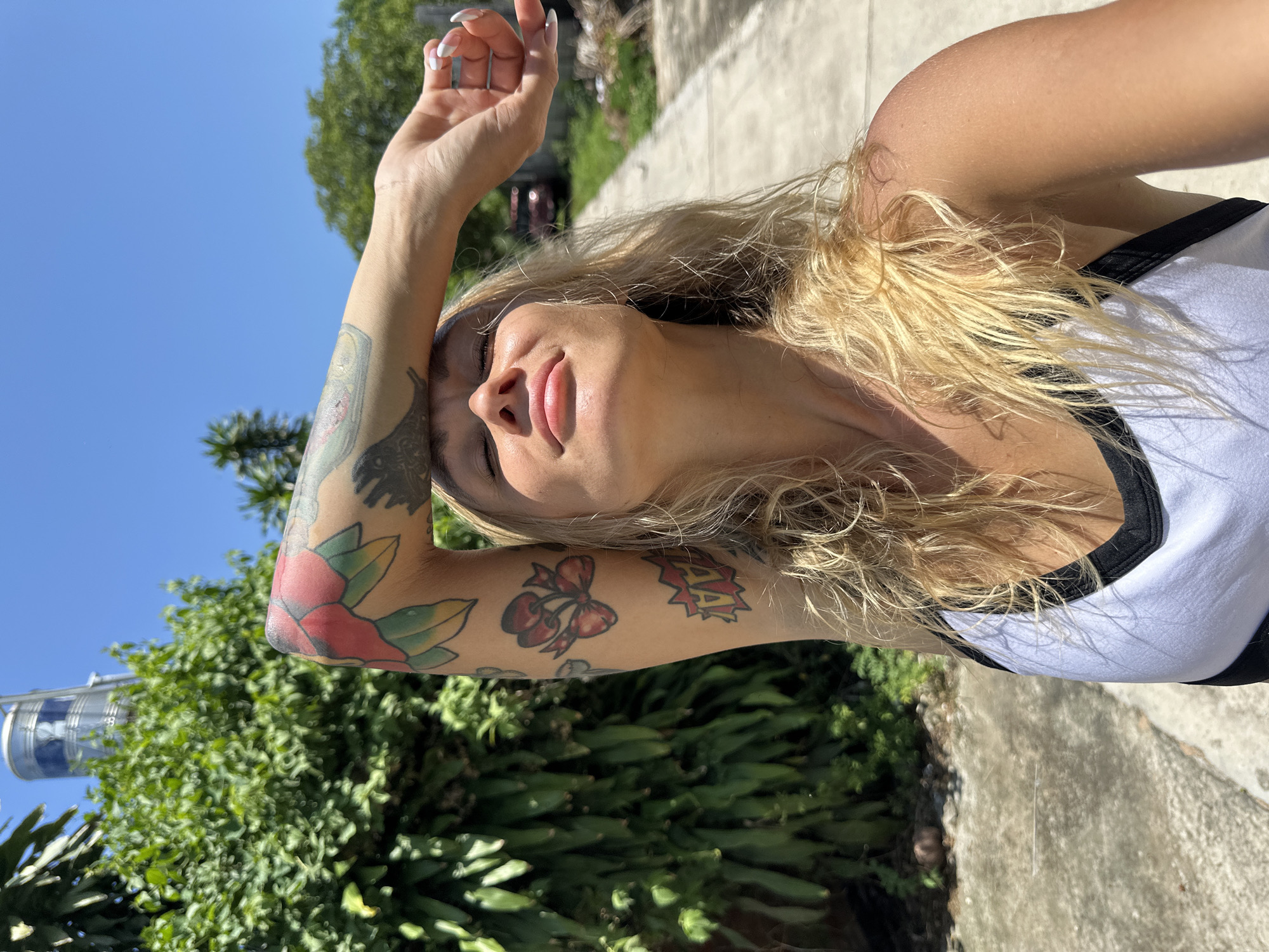 A blonde woman with tattoos on her arm is standing outside in Mũi Né, Vietnam, shielding her eyes from the sun. She is wearing a white tank top with black trim. To her left is lush green foliage, and to her right is a concrete pathway. In the background, a silver water tower is visible against a clear blue sky.