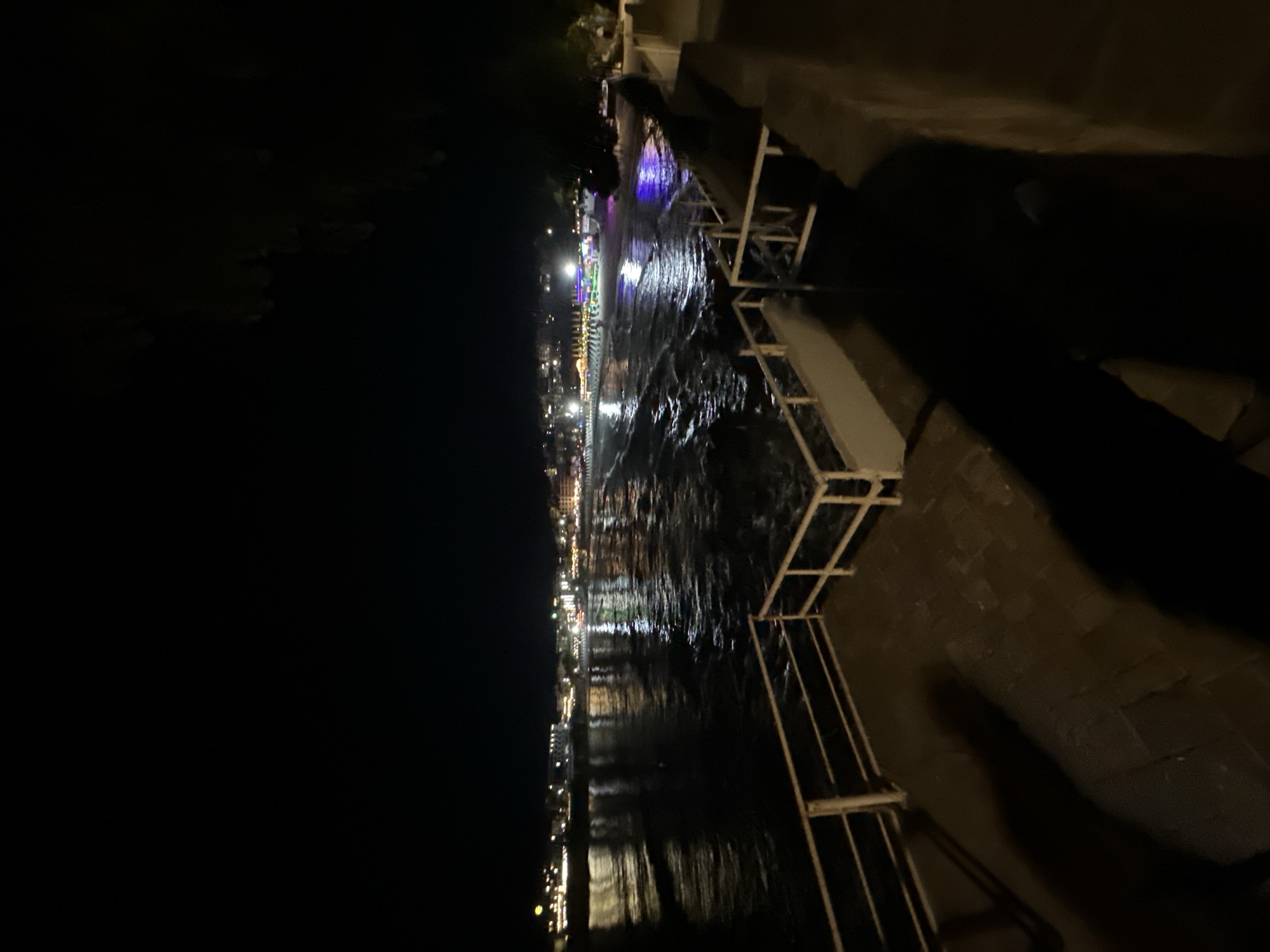 An outdoor, medium shot captured at night shows a body of water reflecting city lights. In the foreground, a brick patio with a shadow of a person is bordered by a white railing. The water in the midground is choppy and reflects the lights of a distant city, which is visible on the horizon. The city appears to be a resort town, with numerous buildings and what looks like beach chairs on the shore. The sky is dark, indicating nighttime.