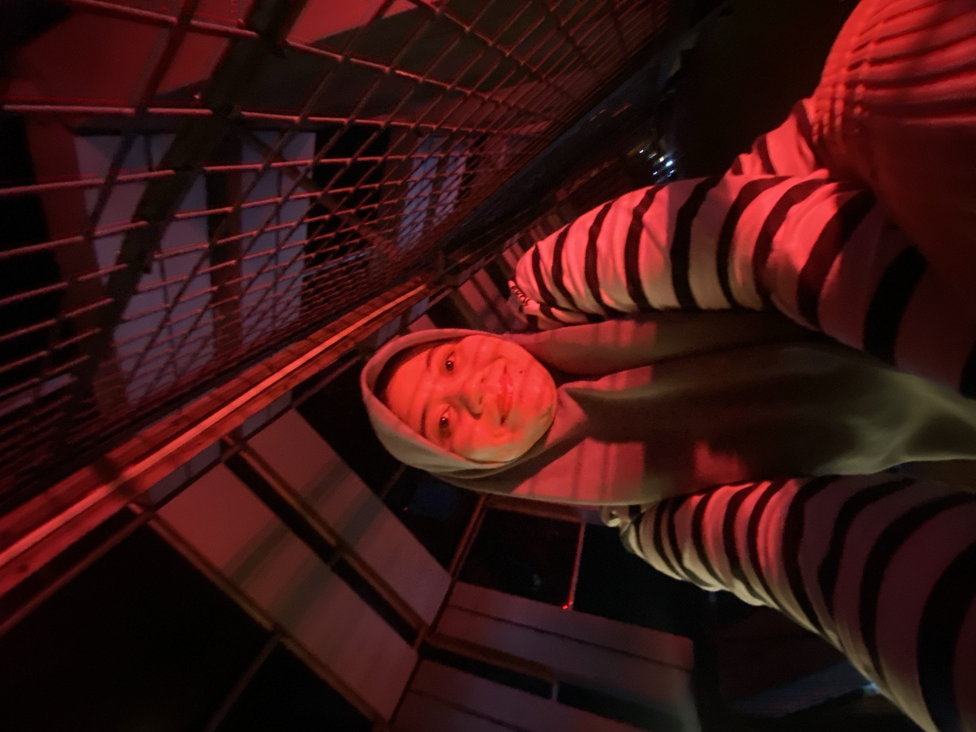 In the city of Banjarbaru, Indonesia, a young woman wearing a grey hijab and a striped shirt is taking a selfie. She is positioned in a structure with a metal grate ceiling illuminated by red lights, creating a dramatic, low-light environment. The background shows angled wooden beams and the dark night sky. The woman's face is partially lit by the red glow, highlighting her smile and expressive eyes.