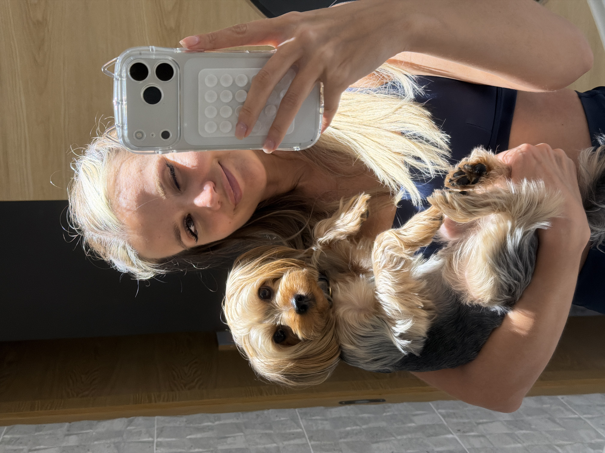In an indoor setting with grey tiled walls and a wooden accent on the left, a blonde woman holding a small Yorkshire Terrier in her arms takes a selfie. The woman is wearing a dark blue sports bra and dark blue leggings, and her hair is partially blonde and partially brown, styled loosely. She is winking at the camera, and the dog is looking forward. The woman is holding a smartphone in her right hand, taking the photo. The overall lighting suggests it is daytime, with sunlight casting shadows on the subjects and the environment. The background is slightly blurred, indicating a shallow depth of field. The location is identified as Thep Krasatti, Thailand.