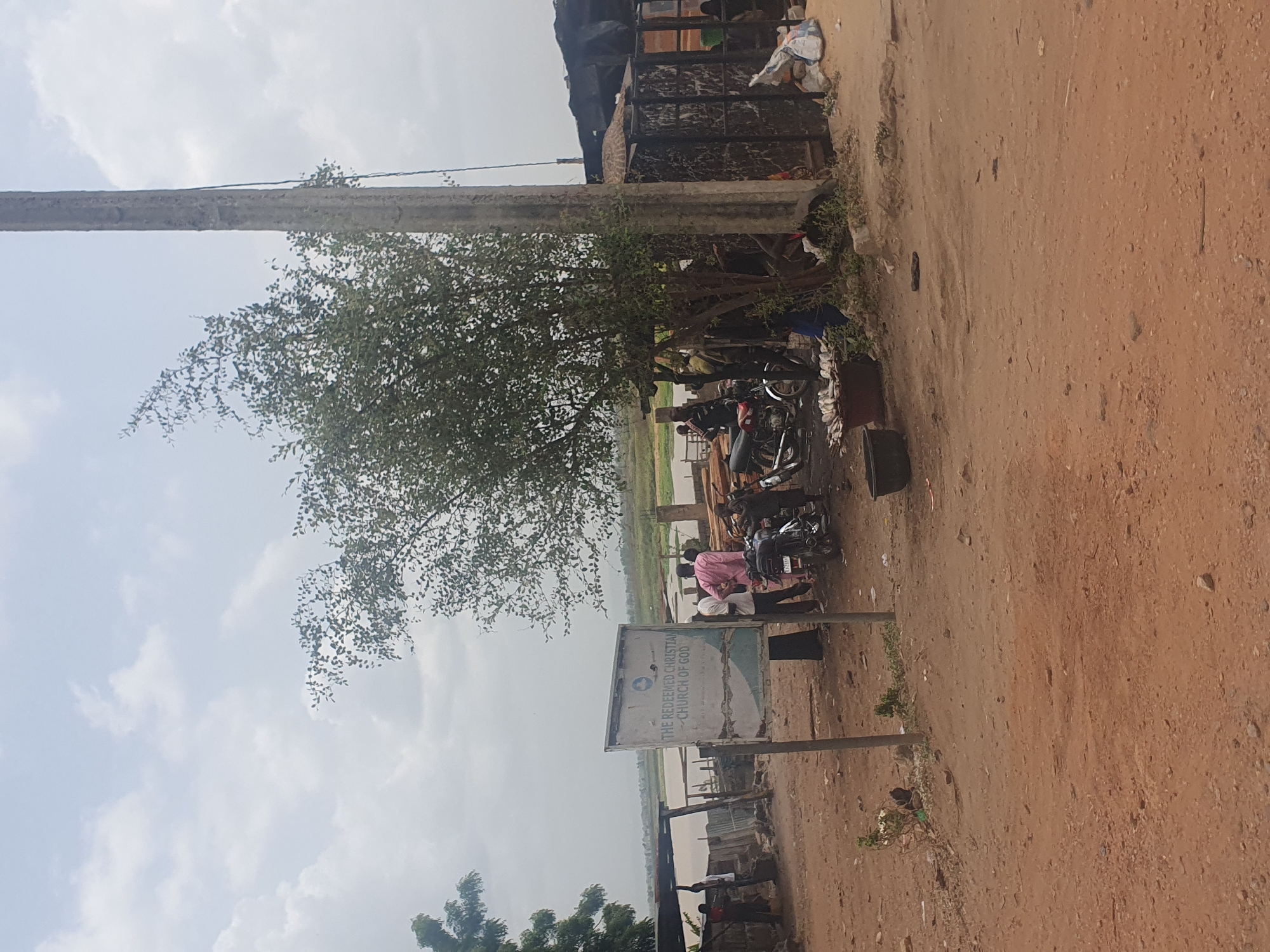 The image captures a scene in Lokoja, Nigeria, near what appears to be a river or body of water. The foreground is dominated by a dirt ground leading to a gathering of individuals. Several motorcycles are parked nearby. A sign for "The Redeemed Christian Church of God" is prominently displayed. A large tree provides shade, and a utility pole is visible in the upper left. Behind the gathering, there is a view of the water, with structures or possibly small boats in the distance. The sky is overcast.