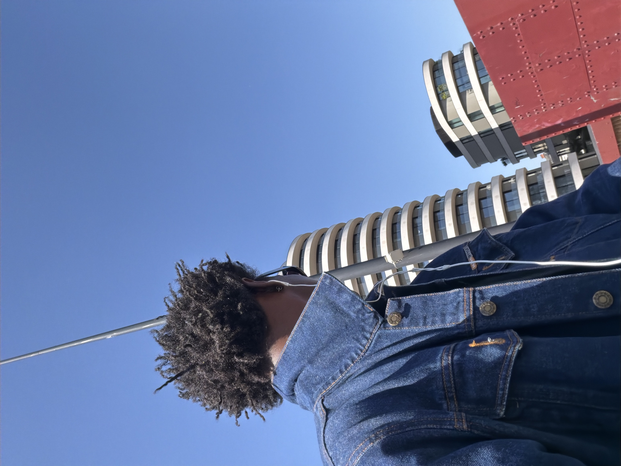 The image captures a low-angle, tilted view of a person looking upwards in a modern urban environment. The main subject is an individual, seen from their right side, whose head, neck, and right shoulder are visible in the lower central part of the frame. They have dark, textured, dreadlocked hair and appear to be wearing white earphones. A blue denim jacket with brass buttons and orange stitching covers their torso.
The background is dominated by a clear, bright blue sky, indicating sunny weather. To the right, modern buildings with distinctive, curved, ribbed facades and glass windows rise upwards, showcasing contemporary architecture. In the upper right corner, a section of a large, red metal structure with visible rivets is prominent, suggesting it could be part of a bridge or a substantial industrial-style building. A slender, metallic bar or pole extends diagonally across the upper left portion of the image, appearing above the person's head.
The perspective is from below, looking up, which accentuates the height of the surrounding structures and the vastness of the sky. The overall scene depicts a moment in a vibrant outdoor setting in London, United Kingdom. No discernible text is visible in the image.