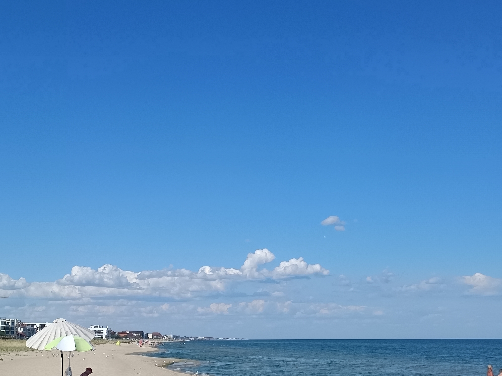 A wide shot captures a serene beach scene under a vast, clear blue sky. Fluffy white clouds drift lazily across the upper portion of the frame, adding texture to the expanse. In the foreground, a sandy beach stretches out, dotted with a few scattered seagulls. A partially open umbrella, adorned with alternating green and white stripes, stands prominently on the sand, its pole casting a subtle shadow. To the left of the umbrella, a person is partially visible, their back to the camera. In the middle ground, the calm, blue ocean meets the shore with gentle waves. Beyond the water, a distant coastline is visible, featuring a line of buildings and what appear to be small structures. The overall atmosphere is peaceful and leisurely, suggestive of a sunny day at the seaside.
