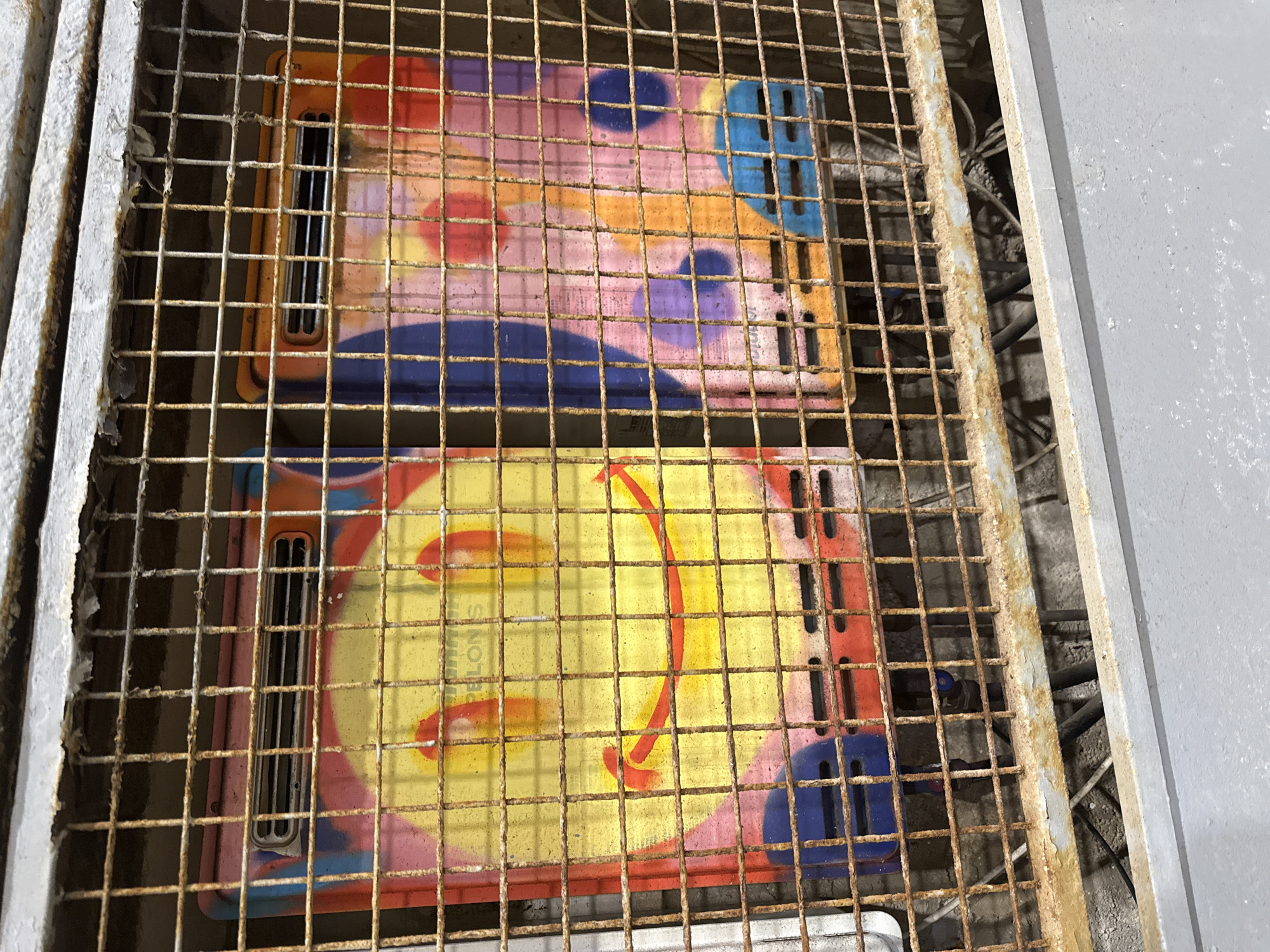 Two colorful rectangular objects are placed side-by-side and are partially obscured by a rusty metal grate. The objects appear to be some kind of panels or covers, possibly for electrical equipment or ventilation, as indicated by the slotted openings on their sides. They are decorated with spray-painted abstract designs featuring various colors, including orange, yellow, pink, purple, and blue. The top object has circular shapes and broad strokes of color. The bottom object has a predominantly yellow background with red curved lines resembling a smiley face or a stylized arrow, along with some pink and blue elements.
The setting appears to be outdoors, given the rust on the metal grate, which suggests exposure to the elements. The grate itself is made of thick metal bars, creating a grid pattern over the objects. To the right of the grate and objects, a gray metallic surface is visible, possibly a wall or a structural component. In the background, behind the objects and grate, a dark, rough concrete surface is visible, and some black cables or pipes can be seen. The lighting suggests daytime, but no specific time is discernible. The location context indicates Tel-Aviv, Israel, but there are no explicit visual cues in the image to confirm this. No people are visible in the frame. There is no clear or legible text visible on the objects.