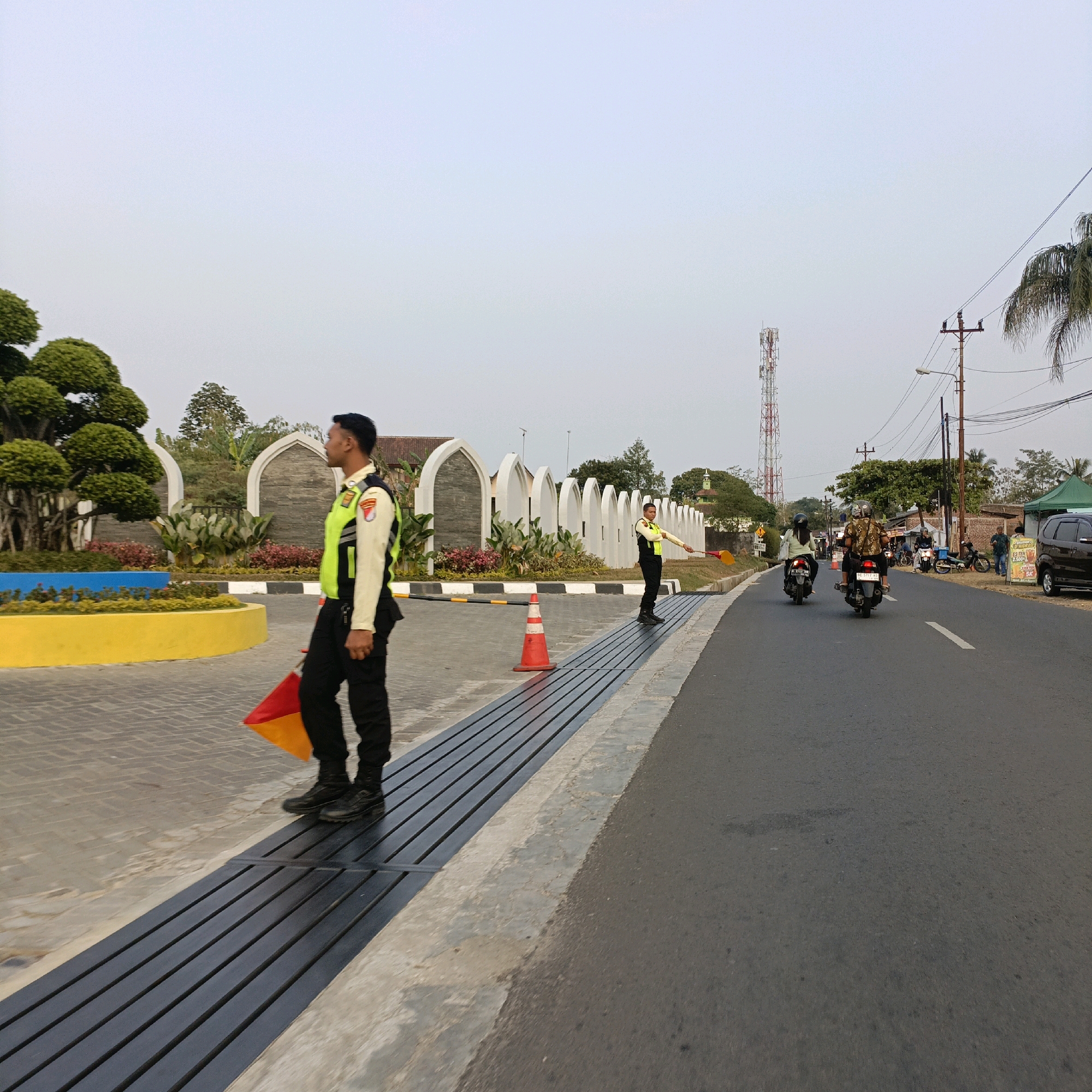 This image depicts a street scene in Salatiga, Indonesia, likely near an entrance or a junction. In the foreground, two individuals wearing high-visibility vests and dark trousers stand by the roadside. The person on the left is facing right, holding a red and yellow flag. The person on the right is facing left, also holding a similar flag. Both individuals appear to be directing traffic or managing pedestrian flow.
The environment includes a paved road with white lane markings, a concrete curb, and a metal grate drainage system running parallel to the sidewalk. On the left side of the road, there is an area with decorative landscaping featuring manicured trees, colorful flower beds (yellow and blue), and white arched structures. In the background, a cell tower is visible against a pale sky, along with power lines and poles. Several motorcycles and a dark-colored car are present on the road, some with riders. On the right side of the road, there is a small stall with a green canopy selling drinks. The overall atmosphere suggests a moderately busy outdoor area.