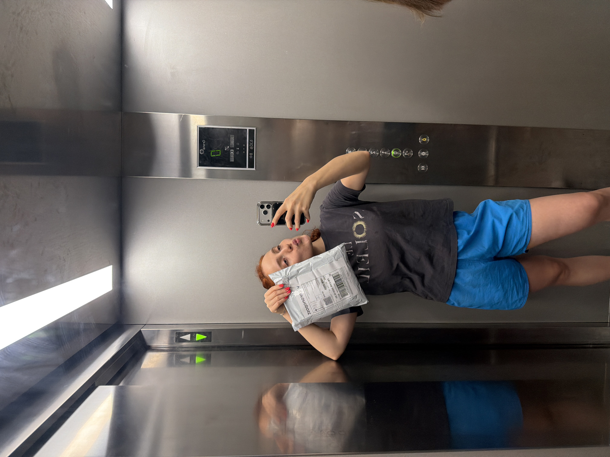 A young woman with red hair is taking a mirror selfie inside an elevator. She is wearing a dark t-shirt with a circular graphic and bright blue shorts. In her right hand, she is holding a small, light-colored package. Her left arm is resting on the elevator wall, and her reflection is visible in the mirrored surface. The elevator has stainless steel walls and illuminated buttons on a control panel to the right of the woman. One of the buttons, displaying a green circle with a "0" inside, is lit, indicating the current floor. The overall environment suggests an indoor, utilitarian space. The location is not specified in the image.
