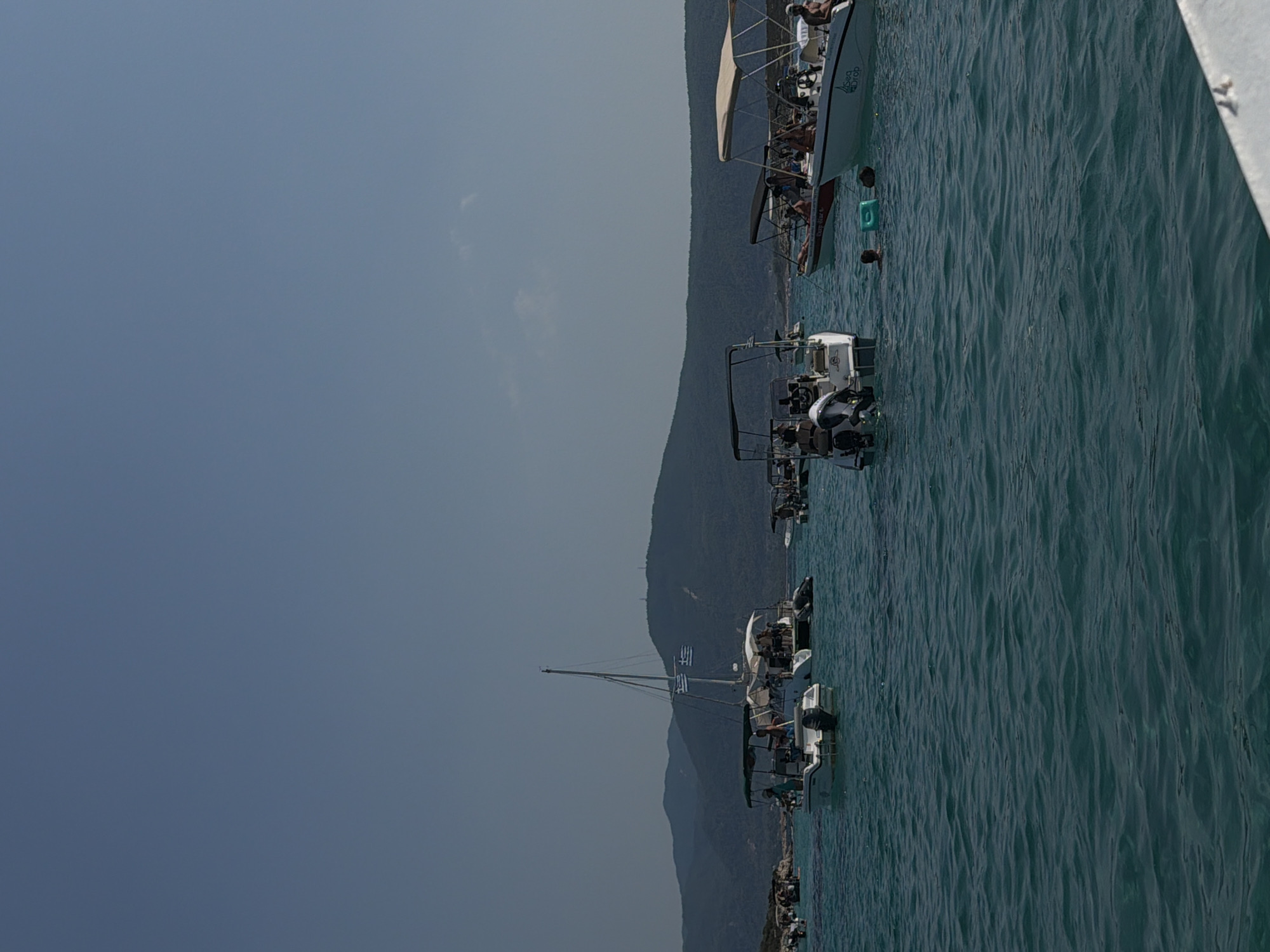 The image depicts a group of boats anchored in the clear turquoise waters off the coast of Greece, under a bright, clear blue sky. In the foreground, the water is rippled and calm. Several small to medium-sized motorboats and a sailboat are visible, with people on board enjoying a day on the water. Some boats are equipped with canopies for shade. In the middle ground, a sailboat with two Greek flags prominently displayed is anchored. Further back, the lush green, mountainous coastline of Greece forms the backdrop, with some visible structures on the hilltops. The scene suggests a popular tourist destination, likely a beach or bay, where people engage in recreational boating and swimming.