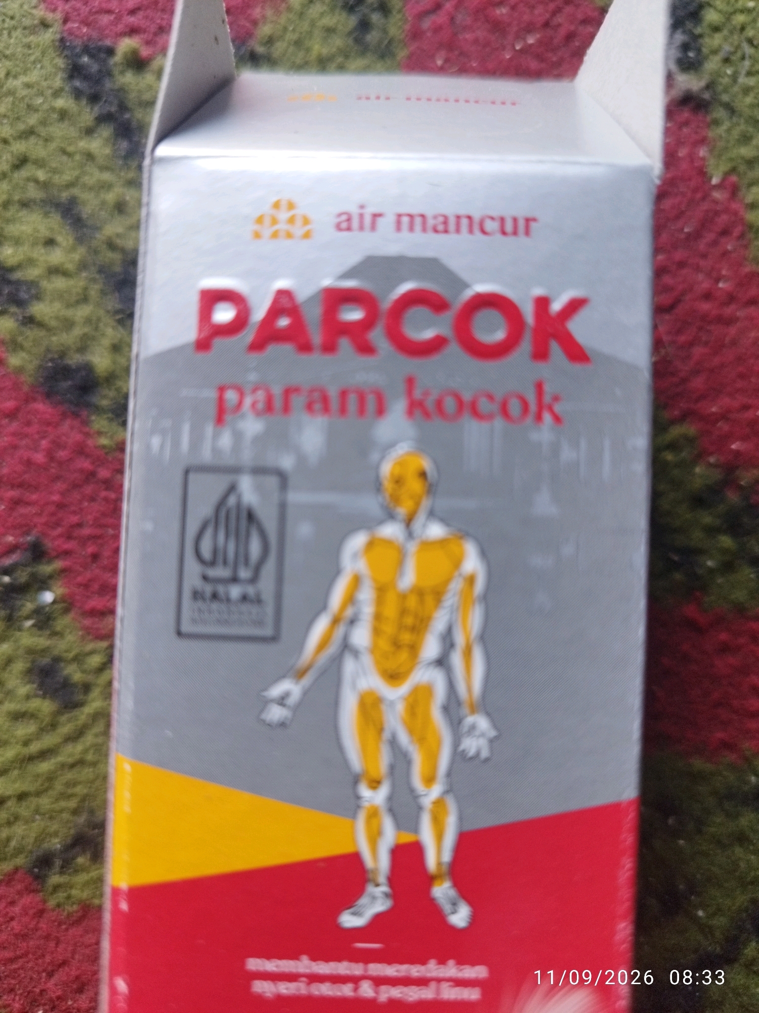 A close-up shot shows a portion of a box of "PARCOK" medicine, a product of "Air Mancur". The box is primarily silver with red and yellow accents. The prominent text on the box reads "PARCOK" in large red letters and "param kocok" in smaller red letters below it. A stylized illustration of the human muscular system in yellow and white is visible on the box. A "HALAL" logo in a gray rectangle is also present. The bottom of the box has red text that is partially obscured. A timestamp of "11/09/2026 08:33" is visible in the lower right corner of the image. The box is placed on a textured surface that appears to be a rug with red and green fibers. There is no discernible information in the image to suggest its location in Mojokerto, Indonesia.