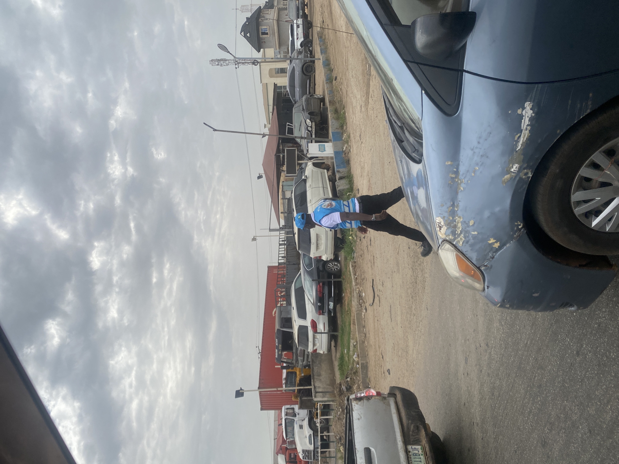 The image shows an overcast street scene in Pakuro, Nigeria. A man wearing a blue and white vest is bent over the front of a blue car with chipped paint. Behind the car, a line of parked vehicles extends into the distance, including cars, vans, and trucks. Buildings and utility poles are visible along the roadside. The sky is filled with grey clouds.