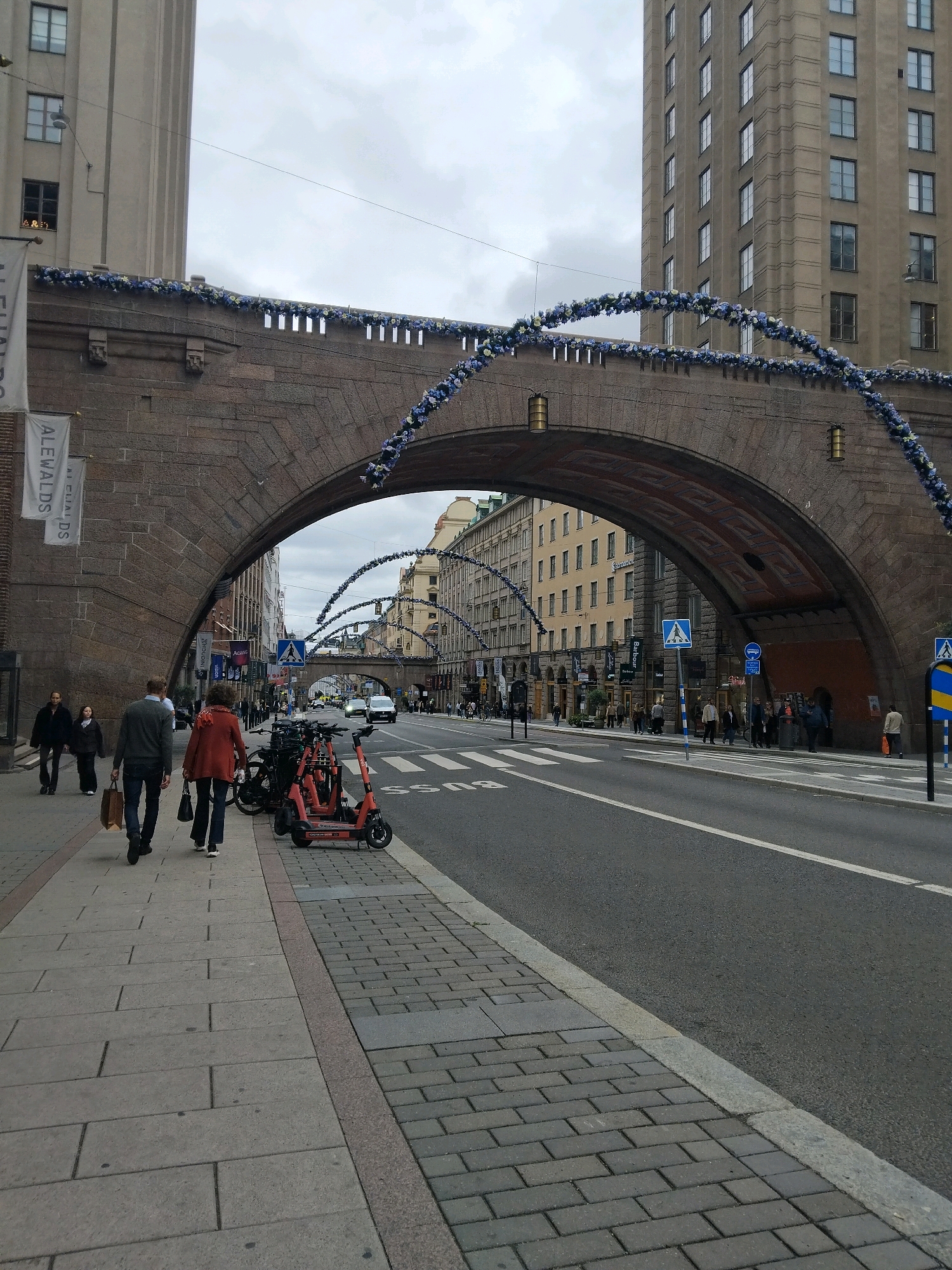 The image shows a street scene in Stockholm, Sweden, under a stone bridge decorated with blue and white garlands. The bridge is an architectural feature of the city, likely a viaduct or part of a larger structure, with an arched opening that allows traffic and pedestrians to pass beneath. On the left side of the image, a sidewalk runs alongside the street, with several people walking. Among them are a man and a woman, who appear to be a couple, and a younger girl walking slightly behind them. The man is wearing a gray sweater and dark pants, and he is carrying a brown paper bag. The woman is wearing a red jacket and dark pants. On the right side of the sidewalk, several electric scooters are parked. Beyond the sidewalk, a street is visible with traffic, including cars and what appears to be a bus. Buildings line the street, with shops and businesses on the ground floor. The architecture of the buildings suggests an urban environment, possibly a historic or commercial district. The sky is overcast, indicating a cloudy day. The overall scene depicts a bustling urban environment in Stockholm, with a blend of pedestrian activity, vehicular traffic, and notable architectural elements.