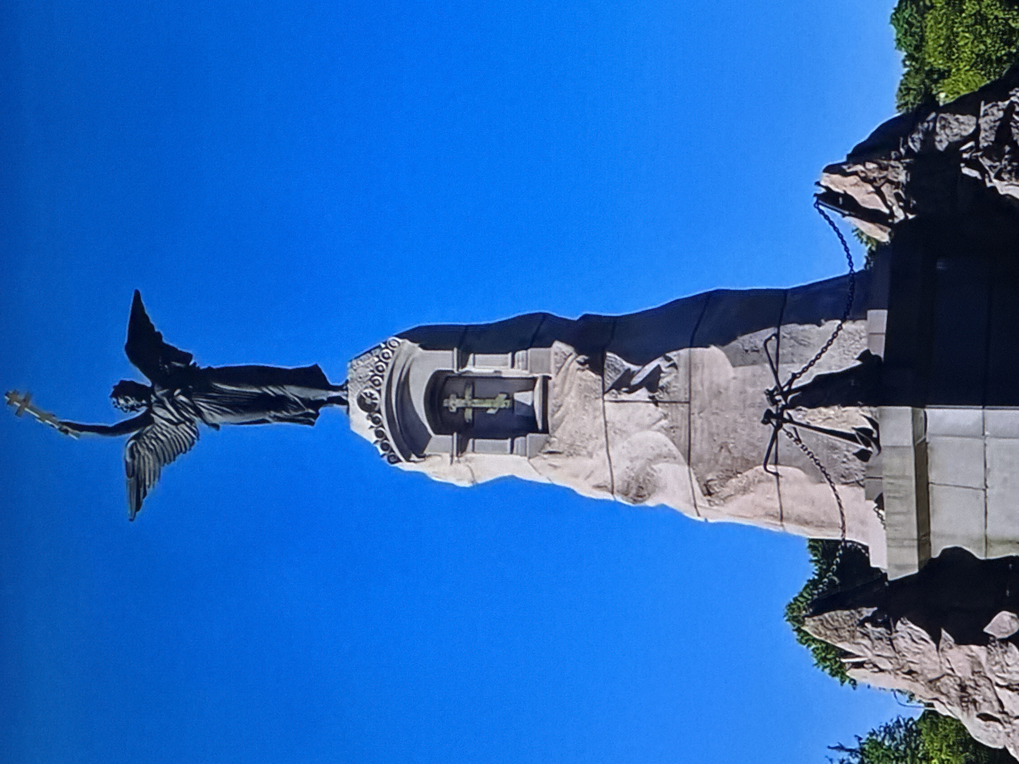 This image shows the Monument to the ”, located in Tallinn, Estonia. The monument features a bronze angel statue with outstretched wings, holding a cross aloft. The angel stands atop a tall, rough-hewn stone pedestal, which is adorned with a niche containing a smaller cross. The base of the monument is surrounded by rocky formations and decorative chains. The sky is a clear, vibrant blue.