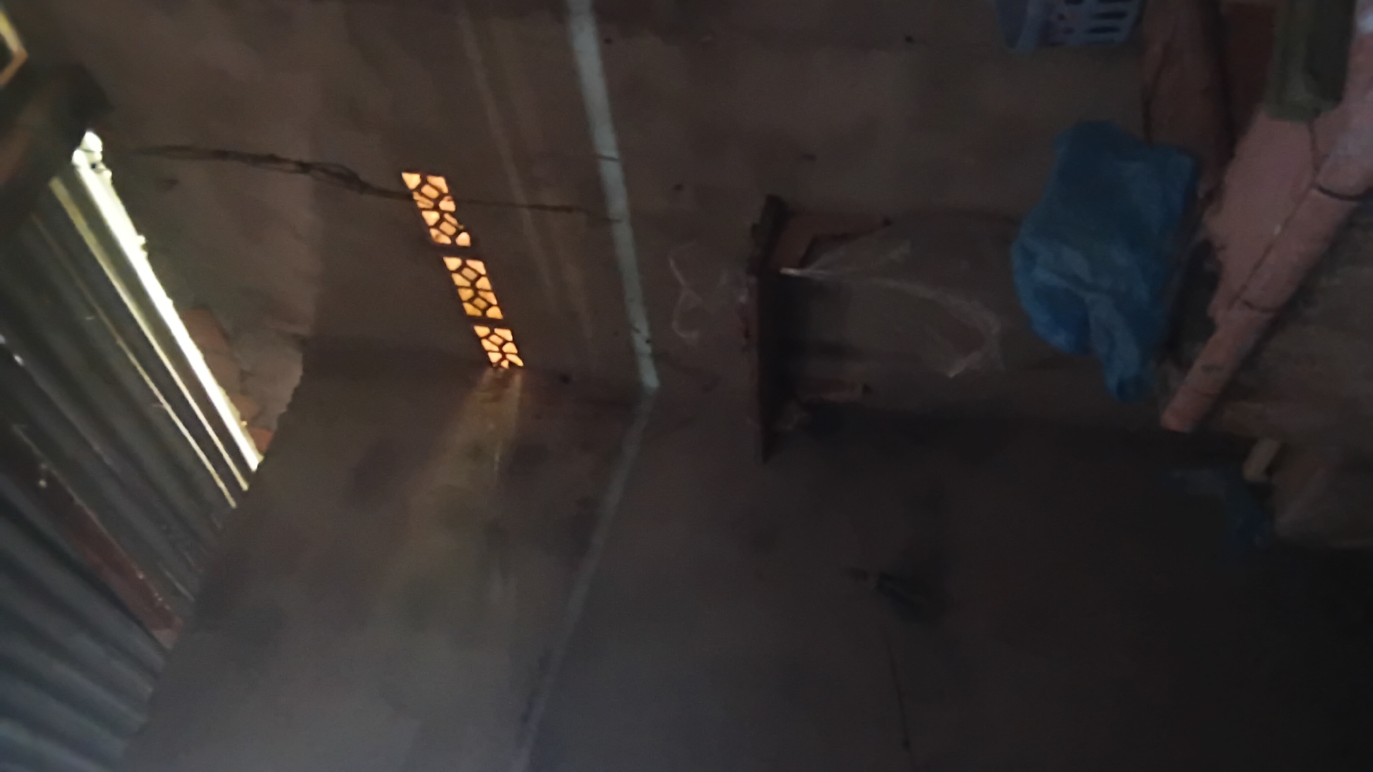 The image depicts the interior of a dimly lit, unfinished room, possibly a storage area or a makeshift shelter, located in Prabumulih, Indonesia. The walls are made of rough, unplastered concrete or mud, and the ceiling is constructed from corrugated metal sheeting, with visible gaps allowing sunlight to stream in. A small, decorative ventilation grate with a geometric pattern is embedded in the upper corner of the wall. A simple wooden shelf is mounted on the wall, holding a few indistinct items, including what appears to be plastic sheeting. In the foreground, on a raised concrete surface, lies a crumpled blue plastic bag and other objects that are not clearly identifiable due to the low lighting and blurriness. To the right, a blue plastic basket is partially visible. The overall impression is one of a rudimentary and incomplete construction.