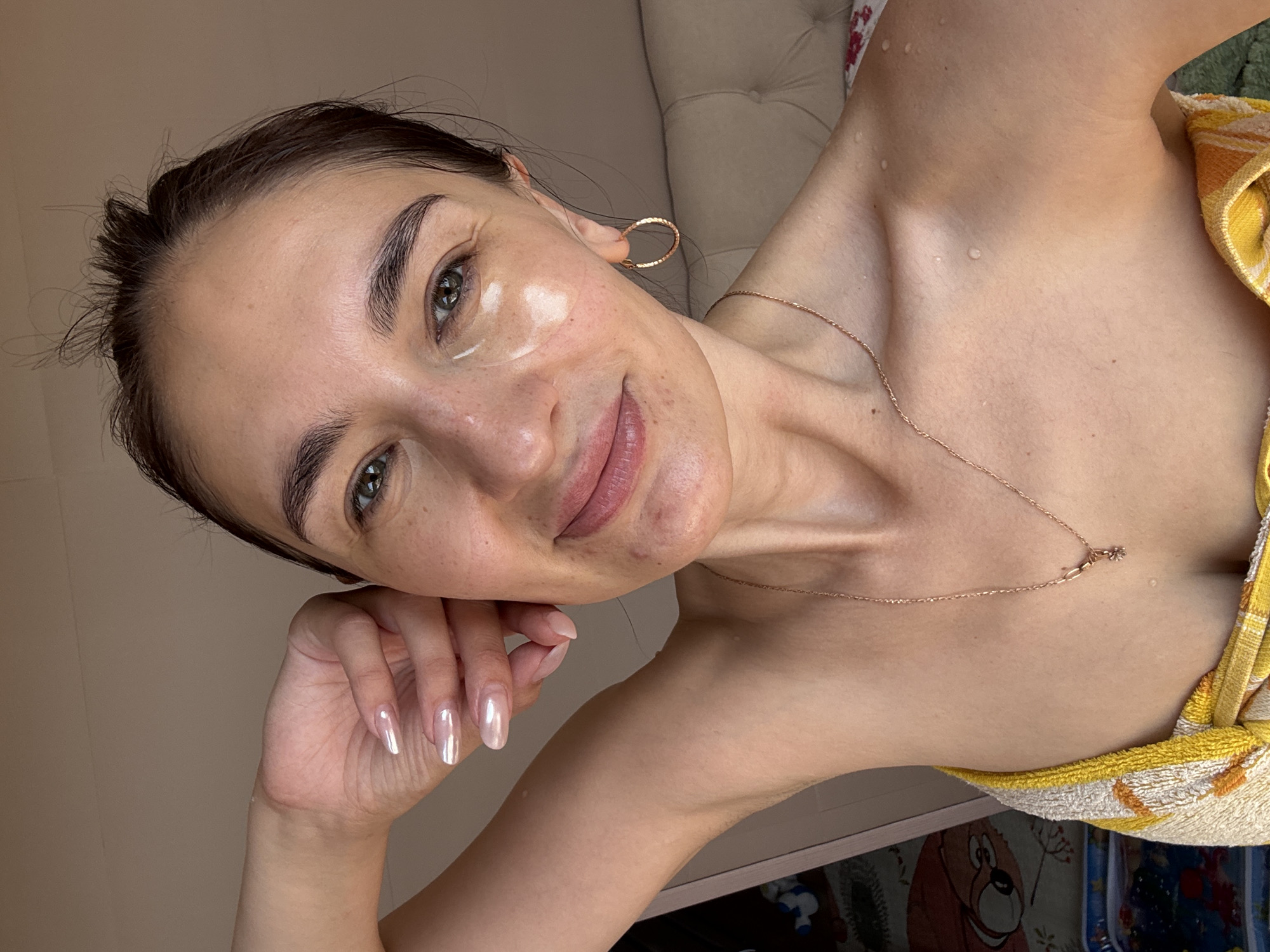 The image shows a woman with fair skin and dark hair pulled back, posing for a selfie. She is wearing a gold necklace and hoop earrings, and her fingernails are long and polished. There is a small, translucent patch on her cheek. Water droplets are visible on her neck and chest, and she is partially covered by a yellow and white towel. The background includes a beige tufted surface and a patterned fabric featuring a cartoon character. There is no evidence to definitively confirm the location as Sochi, Russia.