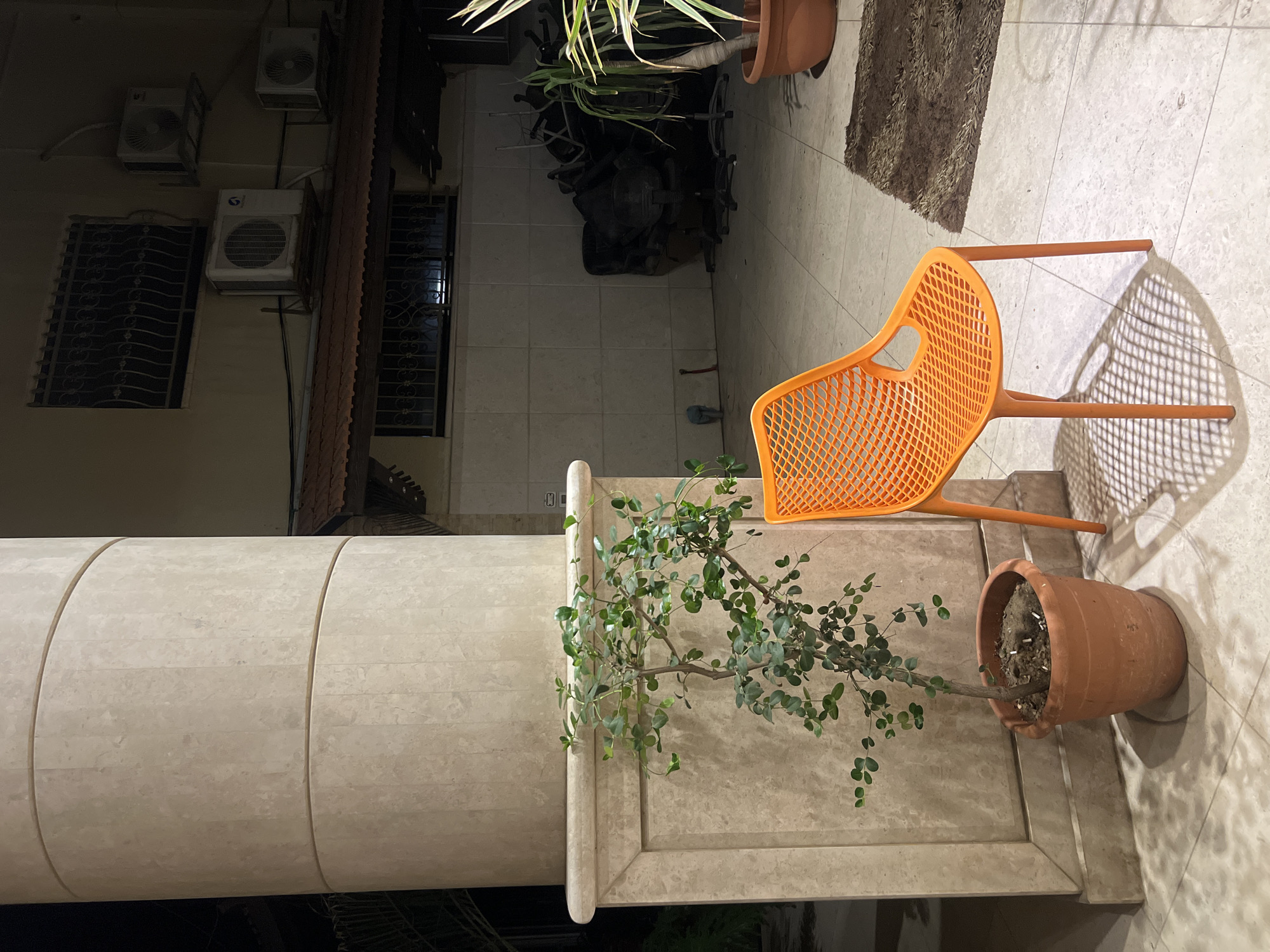 The image depicts an outdoor patio area at night, likely in the city of Gaza, Palestinian Territories. In the foreground, a large, light-colored stone column stands to the left, partially obscuring a potted plant with green leaves. To the right of the plant and column, a bright orange plastic chair with a mesh seat and back is positioned on a tiled floor, casting a patterned shadow. Behind the chair, a dark, shaggy rug is visible. Further in the background, a building with beige tiled walls features several air conditioning units mounted on the exterior. Windows with decorative metal grilles are also present. A pile of office chairs is stacked in the corner of the patio. The scene appears to be a residential or commercial outdoor space, possibly a courtyard or balcony. There are no people visible in the image.