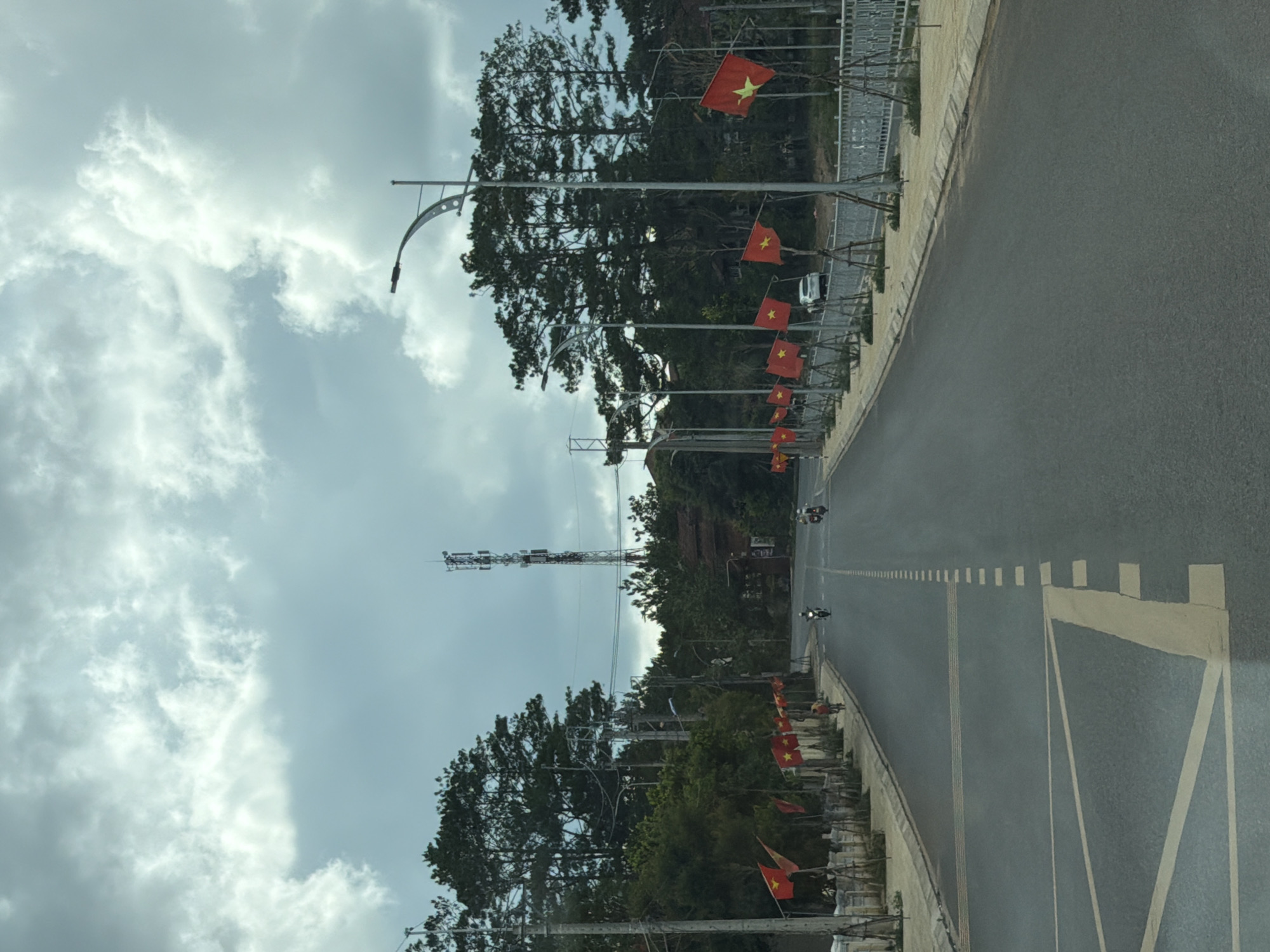 The image captures a wide, paved road in Măng Đen, Vietnam, under a heavily overcast sky. The view is slightly elevated, looking down the length of the road.
**Subjects:** The primary subjects are the road, numerous Vietnamese flags, lampposts, dense green trees, a tall communication tower, and a cloudy sky.
**Scene:** The road is bordered by a concrete sidewalk on the right, which is lined with lampposts. Each lamppost features multiple Vietnamese flags (red with a yellow star) hanging from its pole. On both sides of the road, lush, dark green trees form a dense canopy. A prominent, slender communication tower rises high above the trees on the left side of the frame.
**Objects:** Key objects include the lampposts with their distinct curved arms, the rectangular Vietnamese flags, the multi-tiered communication tower, and painted road markings. The road surface is dark asphalt with various white and yellow lines, including a series of small white rectangular markers leading into the distance and a large, geometric white marking in the foreground.
**People:** Two individuals riding motorbikes are visible on the road in the mid-distance, indicating light traffic.
**Activity or event taking place:** People are traveling on motorbikes along the road. The presence of numerous national flags suggests a display of patriotism or preparation for a national event, or simply a permanent decorative feature.
**Notable details:**
* **Time of day:** Daytime, indicated by the overall brightness, despite the heavy cloud cover.
* **Weather:** Overcast with significant cloud formations.
* **Location cues:** The widespread display of Vietnamese flags definitively places the scene in Vietnam. The communication tower is a prominent landmark.
**Visible text:** No discernible text is visible on the flags or elsewhere in the image.