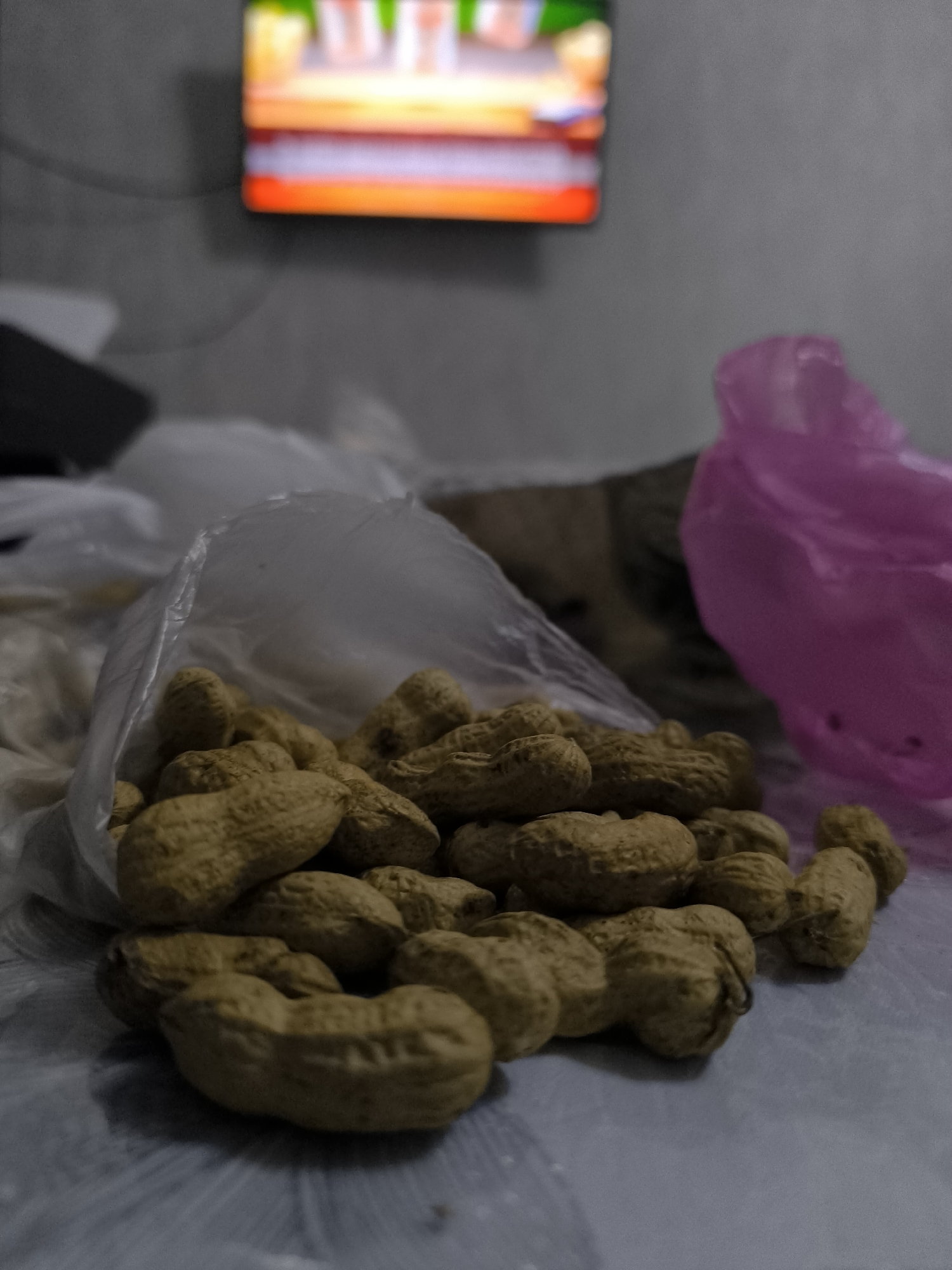 In a dimly lit room, a pile of unshelled peanuts sits in the foreground, spilling out of a clear plastic bag. The peanuts are light brown and textured. Behind the peanuts, a blurry pink plastic bag is visible on the right side of the frame. Further in the background, a television screen displays a colorful, out-of-focus image. The overall environment appears to be a casual setting, possibly a living room or bedroom, with soft surfaces suggesting a bed or couch. The specific location in Kokand, Uzbekistan, is not discernible from the visual cues provided. There are no people or explicit actions depicted, just a still life arrangement of everyday objects.