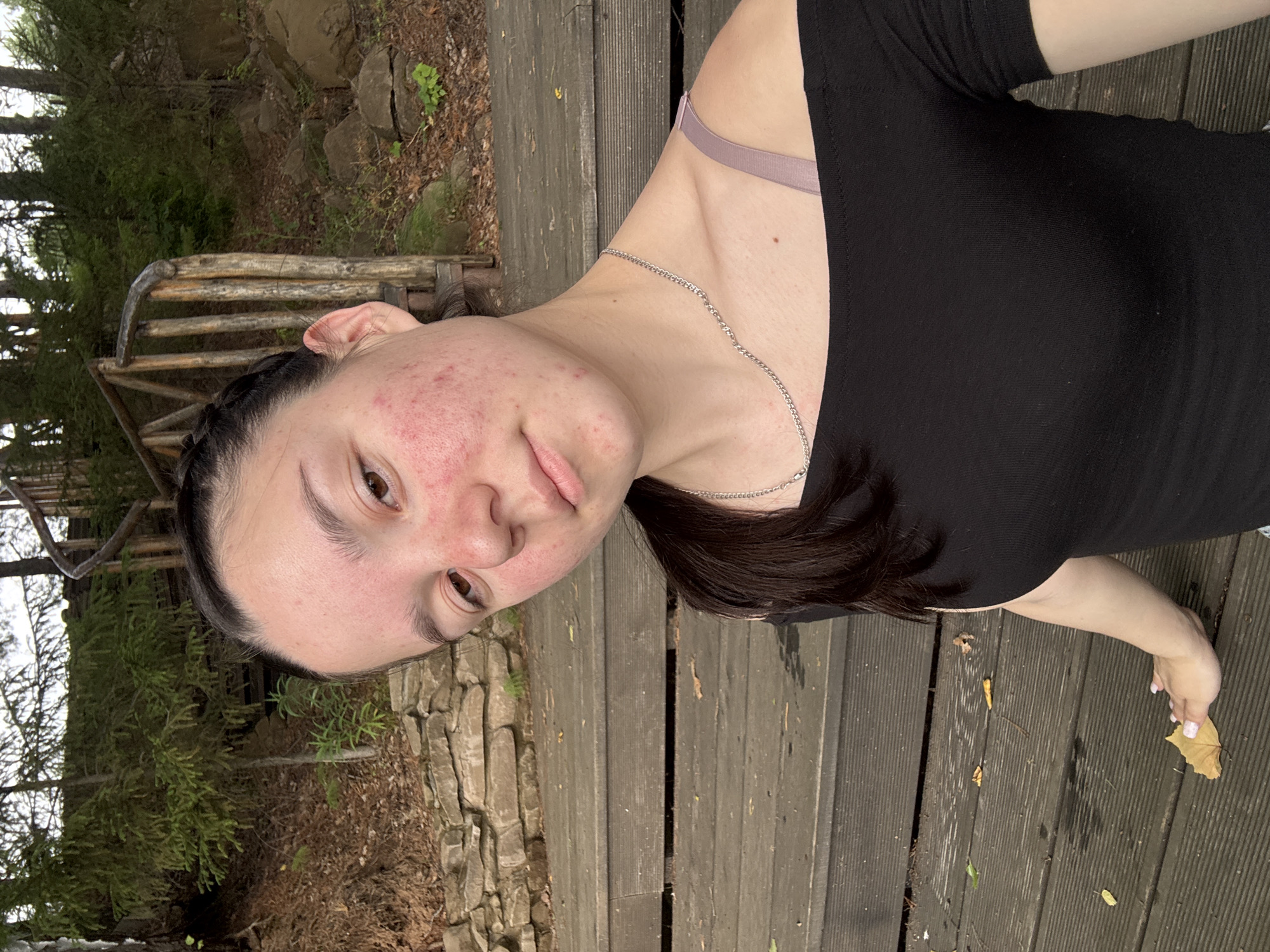 The image shows a young woman with dark hair, appearing to be in her late teens or early twenties, taking a selfie outdoors. She is wearing a black, short-sleeved top and a light-colored bra strap is visible. A silver chain necklace adorns her neck. Her face is turned towards the camera, with a slightly angled pose. Her skin shows some redness and blemishes, particularly on her cheeks. Her eyes are looking directly at the viewer.
The setting appears to be a wooden platform or deck, with a textured, weathered surface. Behind her, to the left, is a rustic wooden railing and a natural landscape with trees, rocks, and fallen leaves. The foliage suggests it might be autumn or a cooler season. The ground is covered with dirt, leaves, and some greenery. The overall lighting suggests it is daytime, possibly overcast given the soft illumination. The woman's expression is neutral or slightly contemplative. Her right hand is visible near the bottom of the frame, gently touching a yellow leaf on the wooden planks. The context provided indicates the location is Pozdyshki, Russia. No text is visible in the image.