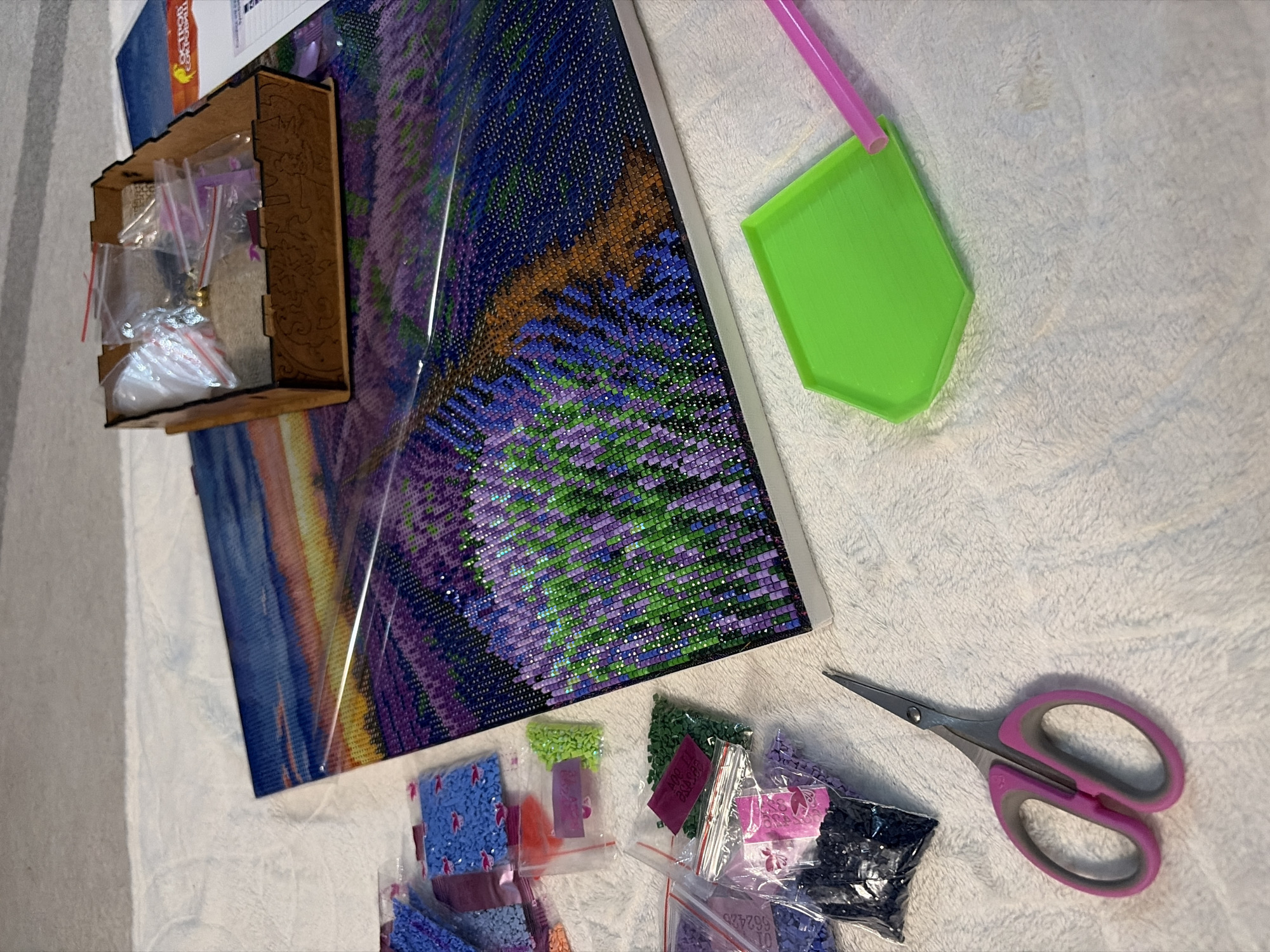 This image shows a diamond painting project in progress. The painting features a field of purple and green, possibly lavender, under a sky with blue and orange hues. The diamond painting is placed on a white, textured surface.
To the left of the painting is a small, wooden storage box containing small, resealable bags filled with various colors of diamond beads. Next to the painting is a green plastic tray and a pink straw. At the bottom of the frame are several resealable bags containing diamond beads in different colors. To the right of these bags is a pair of small scissors with pink handles.
The location is unknown and cannot be determined from the visual information provided. Therefore, information about Chaikovsky, Russia cannot be ascertained.