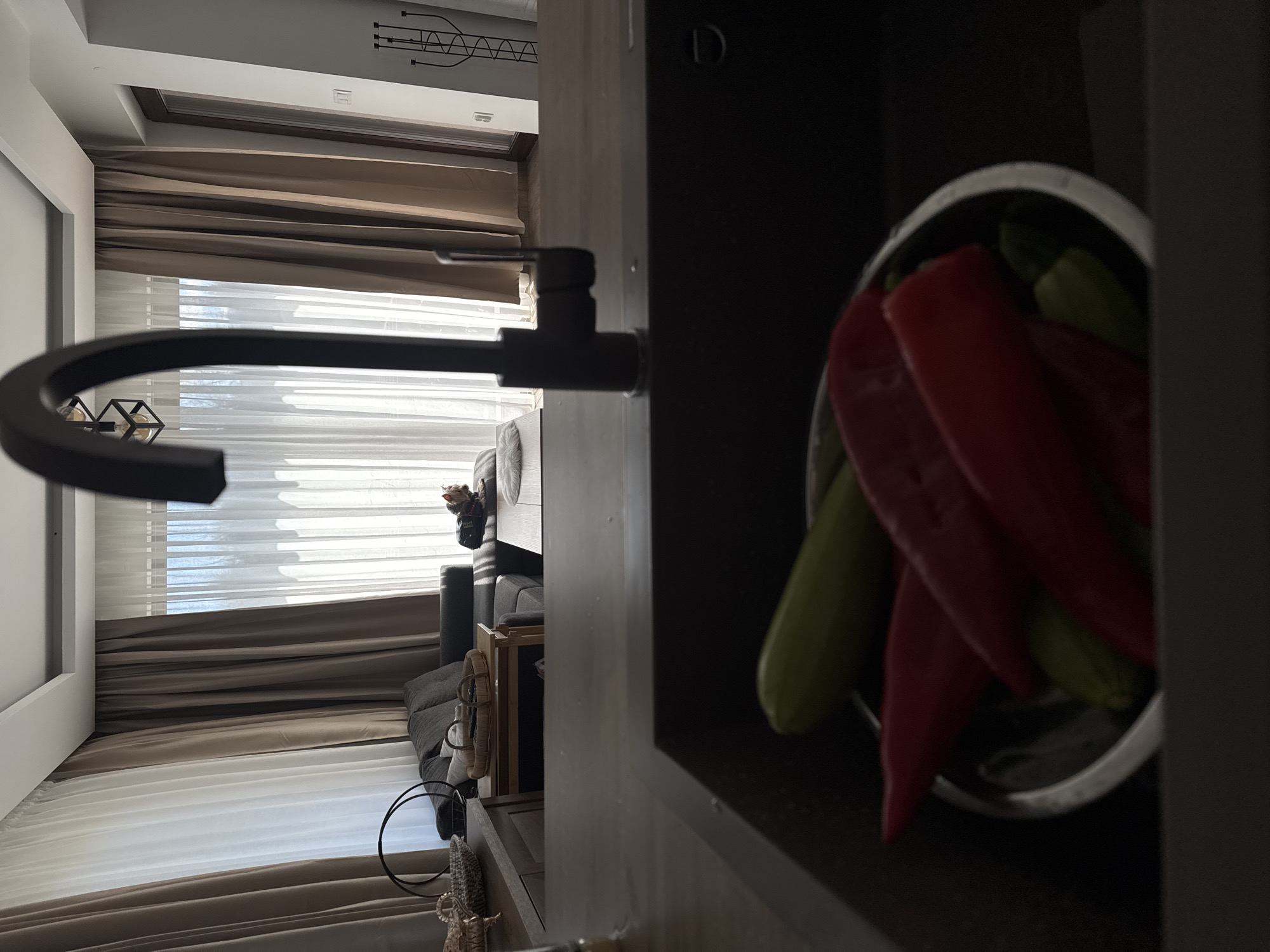 The image shows an interior scene, likely a kitchen or living space. A modern faucet is prominently positioned in the foreground, with a bowl of red peppers and green vegetables visible in the right corner. In the background, a window covered with white and brown curtains provides natural light. A sofa and some furniture are visible behind the faucet, indicating a domestic setting. The location and city are not determinable from the image.