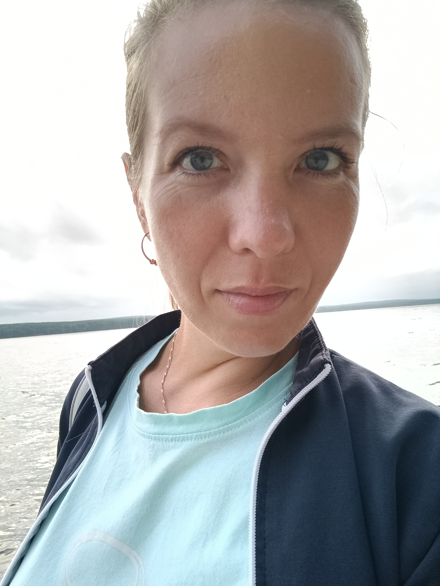 An outdoor, medium close-up selfie shows a Caucasian woman with blonde hair and blue eyes. She is wearing a light blue t-shirt and a dark blue zip-up jacket. A thin, rose gold chain necklace is visible around her neck. She is wearing a small, rose gold hoop earring in her left ear. The background consists of a body of water and a treeline under a cloudy sky. The image appears to have been taken near the Kama River or the Votkinskoye Reservoir, in or near the city of Chaikovsky, Russia.