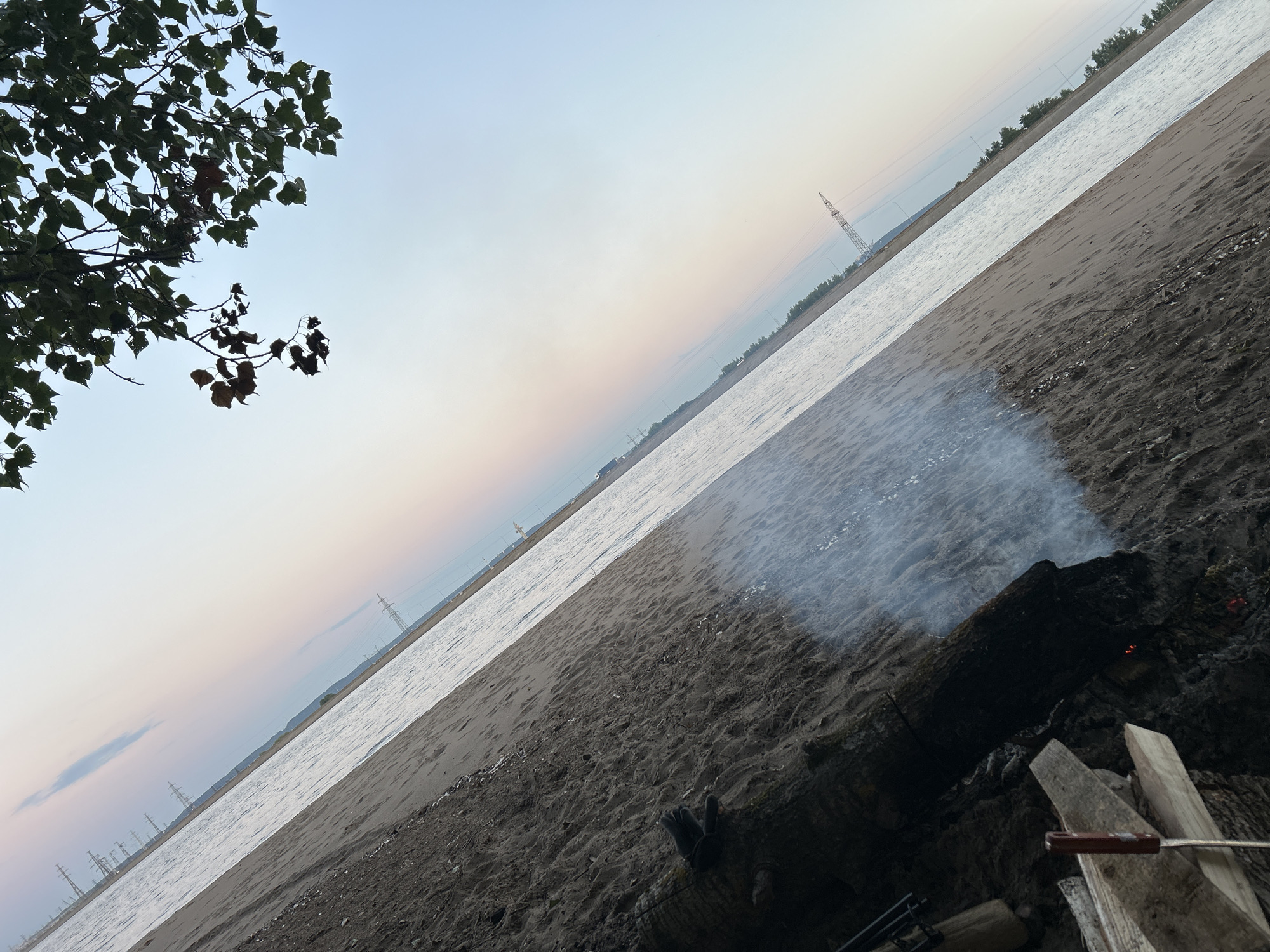 This image depicts a scene on the sandy bank of a wide river, likely the Volga River, near the city of Noviy, Russia, during twilight. The sky displays a soft gradient of pastel colors, transitioning from pale blue at the top to hints of pink and peach near the horizon. In the foreground, a campfire is burning, with smoke gently rising into the air. Several pieces of wood are arranged around the fire, and what appears to be a metal poker with a red handle rests on a piece of lumber. On the sand, to the right of the campfire, a dark-colored glove with two fingers extended is visible. The river occupies the middle ground, its surface reflecting the muted sky. In the distance, along the opposite bank, a treeline is discernible, interspersed with tall, slender power line towers that stretch towards the sky. The overall impression is one of a tranquil, natural setting at the close of day.