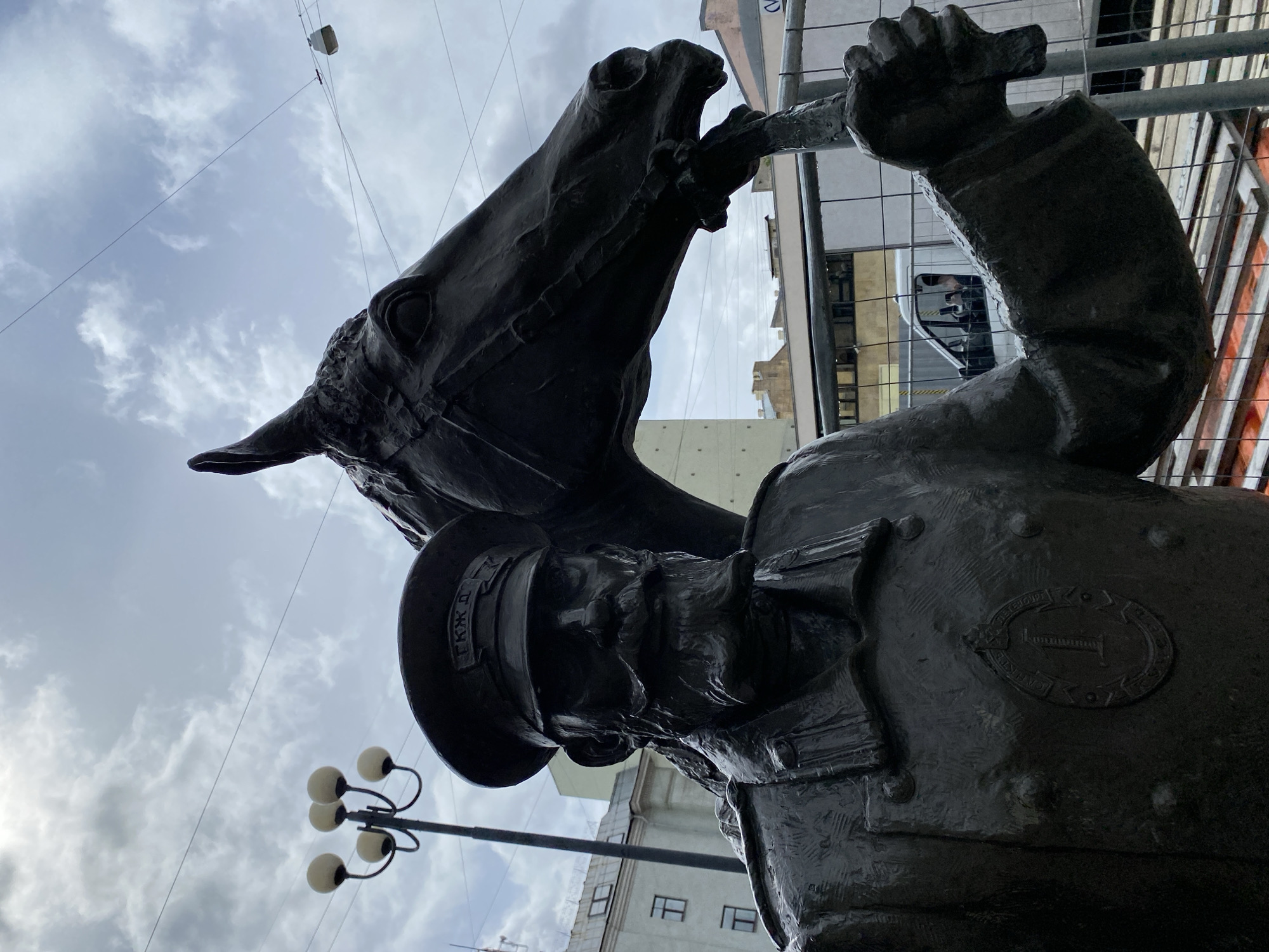 The image shows a close-up of the bronze equestrian statue of Yakov Petrovich Baklanov in Saint Petersburg, Russia. Baklanov is depicted in military uniform and hat. A horse is rearing up with its mouth open, appearing to be holding a trumpet. The statue is set against a backdrop of buildings and a cloudy sky. There is also a street light with round bulbs. Construction materials are partially visible in the background.