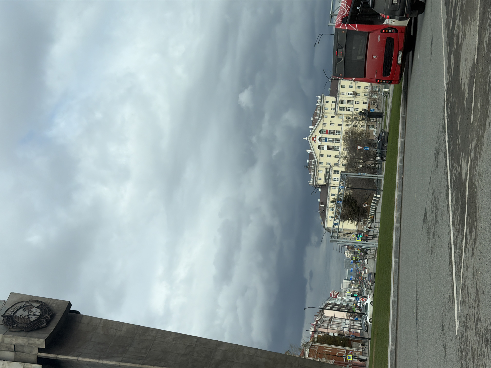 The image depicts a street scene in the city of Perm, Russia, under a cloudy sky. In the foreground on the left is part of a grey monument. A red bus is parked or moving along the right side of the road. The asphalt is wet, suggesting it may have recently rained. A grassy median divides the road. Buildings of varying heights and architectural styles line the street, extending into the distance. Traffic lights are visible, and other vehicles can be seen farther down the street. The sky is filled with heavy, grey clouds.