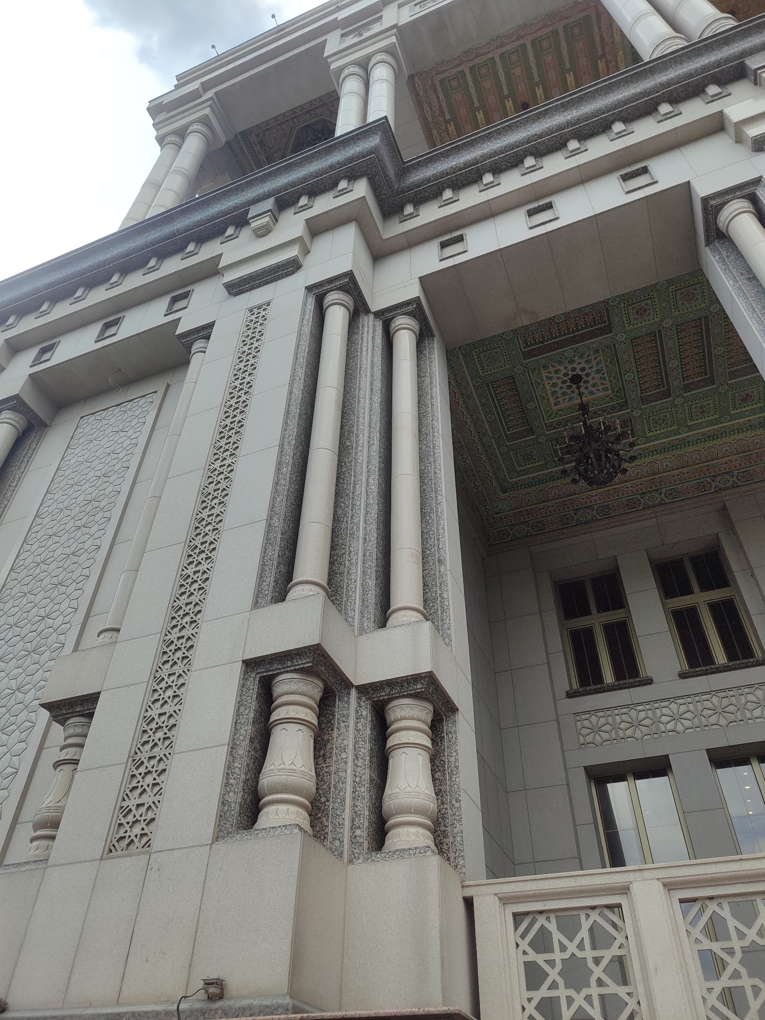 An exterior low-angle shot captures the ornate architectural details of a light-colored building in Dushanbe, Tajikistan. The building features multiple levels with tall, fluted columns and decorative stone carvings. The facade is adorned with intricate geometric patterns on both large panels and narrow vertical strips. Above the columns, the building's upper stories are visible, showcasing windows with dark frames and a highly decorated ceiling with colorful patterns and a chandelier. The sky is overcast and visible in the upper left corner. The image is focused on the building's facade, with no people or other distinguishing elements visible. The building's style suggests it might be a mosque or a government building.