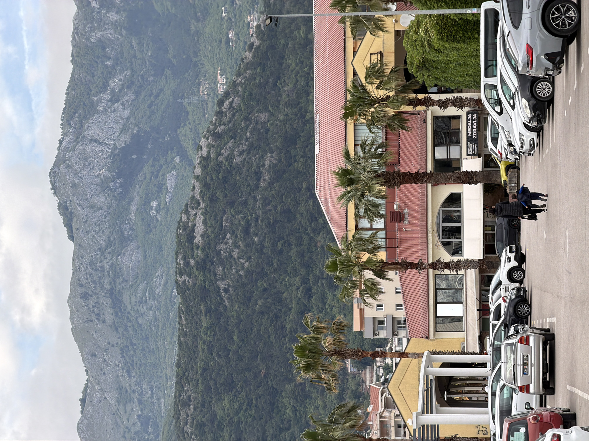 The image shows a street in Budva, Montenegro, with a large mountain range in the background. The right side of the image features a row of parked cars along the street. Buildings with red roofs and palm trees line the street, separating the road from the mountain range. Two people walk on the sidewalk between the cars. The weather appears overcast.