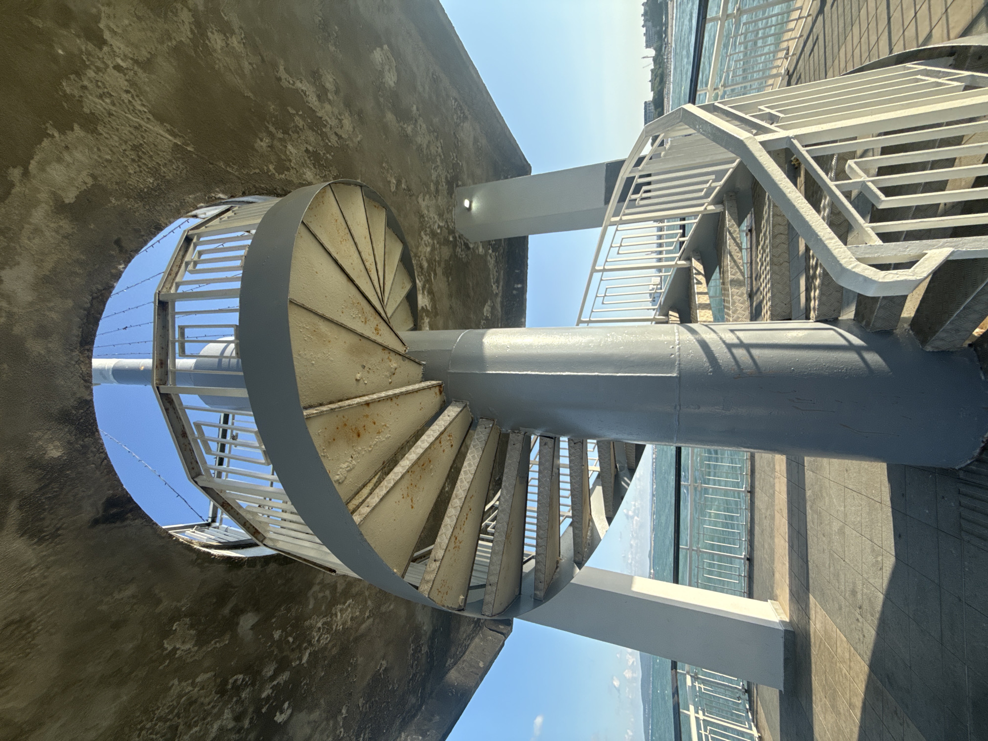 The image shows a spiral staircase leading upwards, situated under a large, textured concrete overhang. The staircase is made of grey metal, with metal treads and railings. The central support column of the staircase is also grey metal. There are white metal railings along the top of the staircase, and further down, a white metal railing outlines a walkway or platform. The walkway is paved with square tiles and is shaded in places by the structure above.
In the background, a body of water, likely the sea, is visible with gentle waves. A coastline with buildings and structures can be seen in the distance under a clear blue sky. The overall impression is of an outdoor public space, possibly a pier or a viewing platform.
This spiral staircase is located at the Burgas Sea Station in Burgas, Bulgaria. It's part of the structure that allows access to the upper levels of the station, offering views of the sea and the city. The architecture is functional and modern, designed to withstand the coastal environment. There are no people visible in the image.