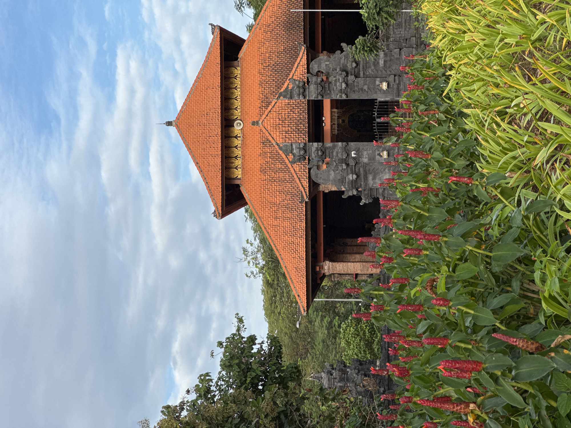 The image presents a view of a structure, likely a religious building, in Kuta Selatan, Indonesia. The building features a multi-tiered orange tile roof. The roof is adorned with a spire at its peak and a decorative gold-colored band along its edge. The building appears to have dark grey stone elements at its entrance, suggesting a Balinese architectural style. Dense greenery surrounds the building, including plants with vibrant red, cone-shaped flowers in the foreground. The sky is partly cloudy with a mix of blue patches and scattered white clouds.