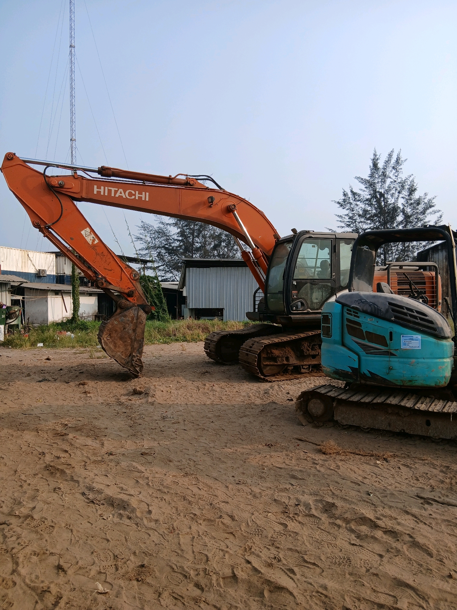 The image depicts an outdoor scene in the city of Pangkalpinang, Indonesia, featuring two excavators. The larger excavator, a Hitachi, is orange and positioned on the left side of the frame. Its arm is extended, with the bucket hanging down. Behind the Hitachi excavator, a smaller, teal-colored excavator is visible. Both machines are situated on sandy ground, which is marked with numerous footprints. In the background, there are several industrial-style buildings with corrugated metal siding and some smaller structures. A tall communication tower stands prominently in the far left background. The sky is clear and pale blue. The environment suggests a construction or storage yard.