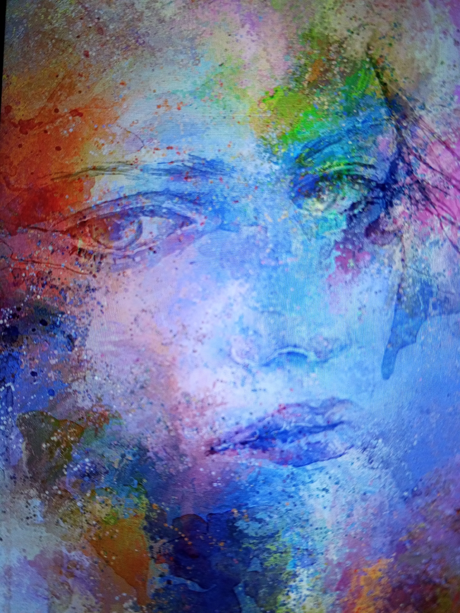 The image features an abstract and vibrant portrait of a human face, rendered in a style reminiscent of watercolor or paint splatters. The face, likely female, is composed of a rich palette of colors including dominant blues and purples, complemented by splashes of orange, red, green, and yellow.
Distinct facial features are visible: two prominent eyes gaze towards the left side of the frame, with the left eye (viewer's left) showing clearer definition in shades of brown and orange. The nose is subtly highlighted by lighter blue and white tones in the center of the face, and the lips are depicted in deeper shades of blue and purple. Streaks of color, possibly representing hair or artistic brushstrokes, flow down the right side of the face (viewer's right).
The entire composition is characterized by a dynamic interplay of color washes, drips, and fine splatters, creating a textured and expressive visual. There is no clear background; instead, the colors and abstract forms seamlessly merge with and surround the facial features. The overall mood conveyed is ethereal or contemplative, with no specific activity taking place. The image appears to be a photograph of a digital display or a print, evidenced by a subtle pixelated grid pattern. No text is visible within the artwork itself. This artwork is associated with Tinaquillo, Venezuela.