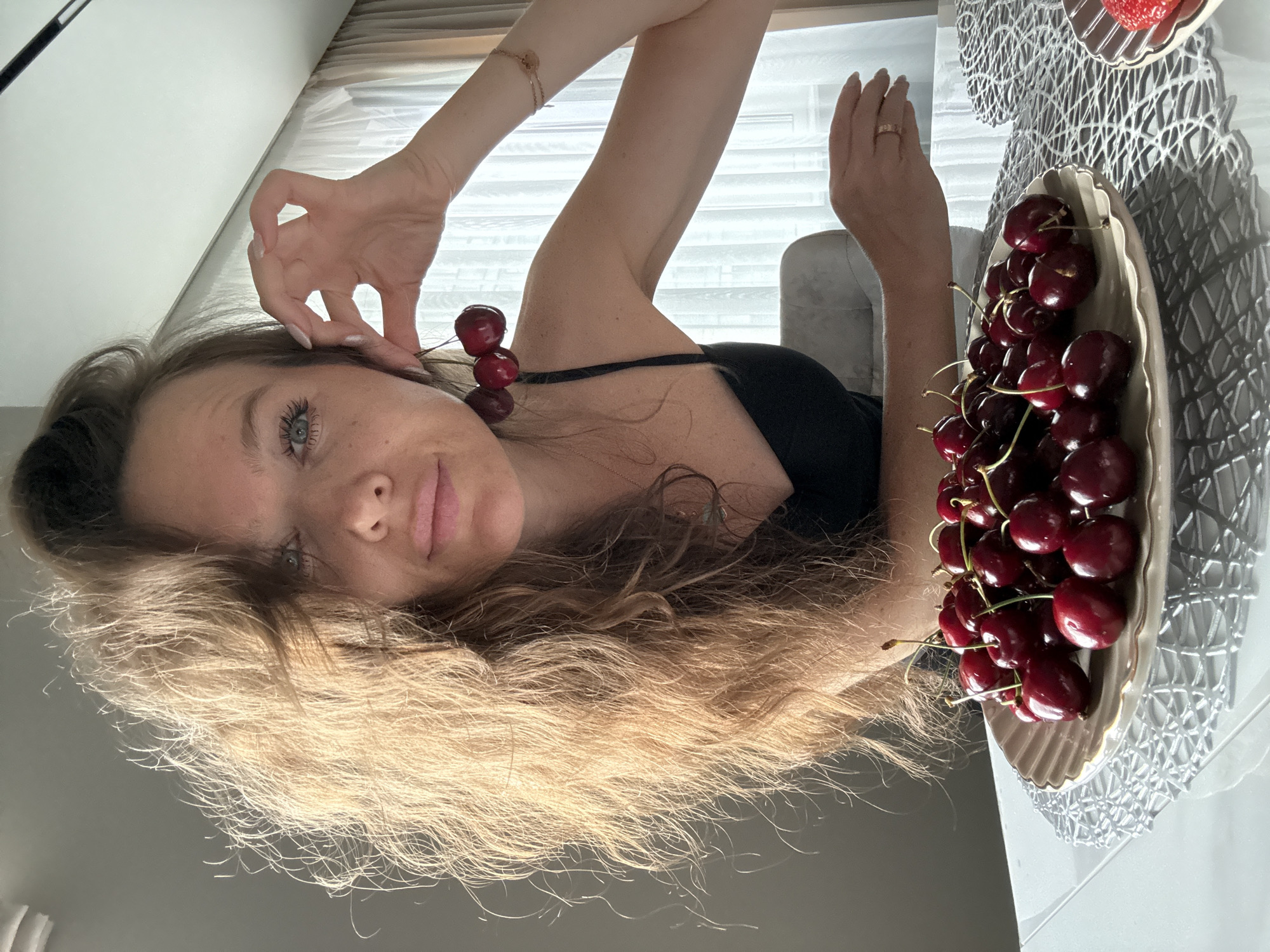 A woman with long blonde hair is lying down, her head resting on her hair. She is looking towards the camera with blue eyes and a neutral expression. She is holding two cherries near her ear. The woman is wearing a black top. In the background, there is a table with a bowl of cherries and a strawberry on a decorative placemat. The setting appears to be indoors, possibly a home, with sunlight coming through blinds in the background. The overall impression is one of relaxation and enjoyment of fruit.