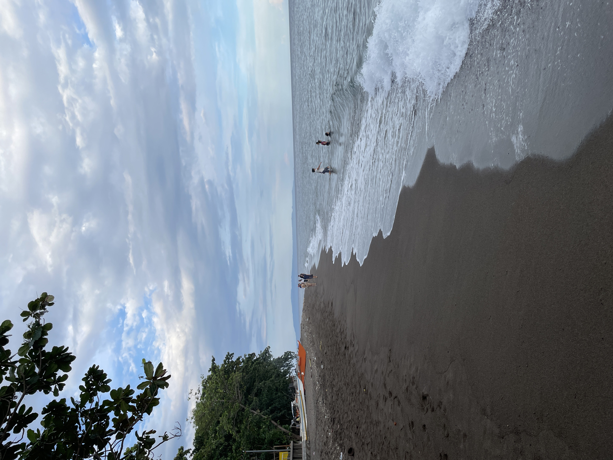 A wide-angle, vertical shot captures a tranquil beach scene in Dauin, Philippines, under a cloudy sky. The dark, volcanic sand of the beach stretches into the distance, marked by footprints and some debris. Several boats are pulled ashore near a cluster of trees on the left side of the frame. In the water, a few individuals are wading and swimming, while gentle waves break on the shore. The horizon is visible in the background, with a distant landmass under the vast expanse of clouds.